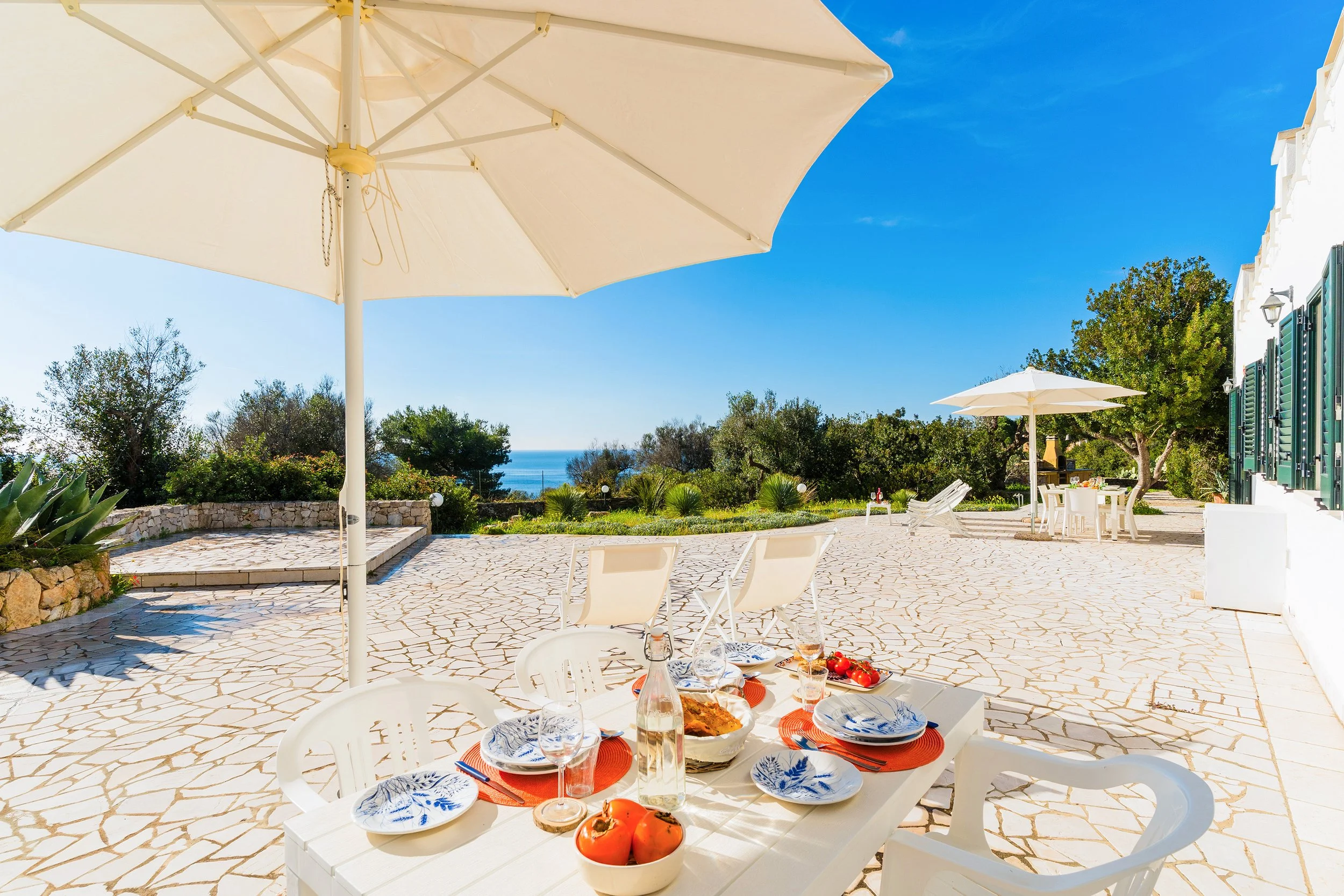An outdoor patio with large white umbrellas, white chairs, and a table set with plates, glasses, and a bowl of tomatoes, overlooking a garden with trees and a distant view of the ocean under a clear blue sky.