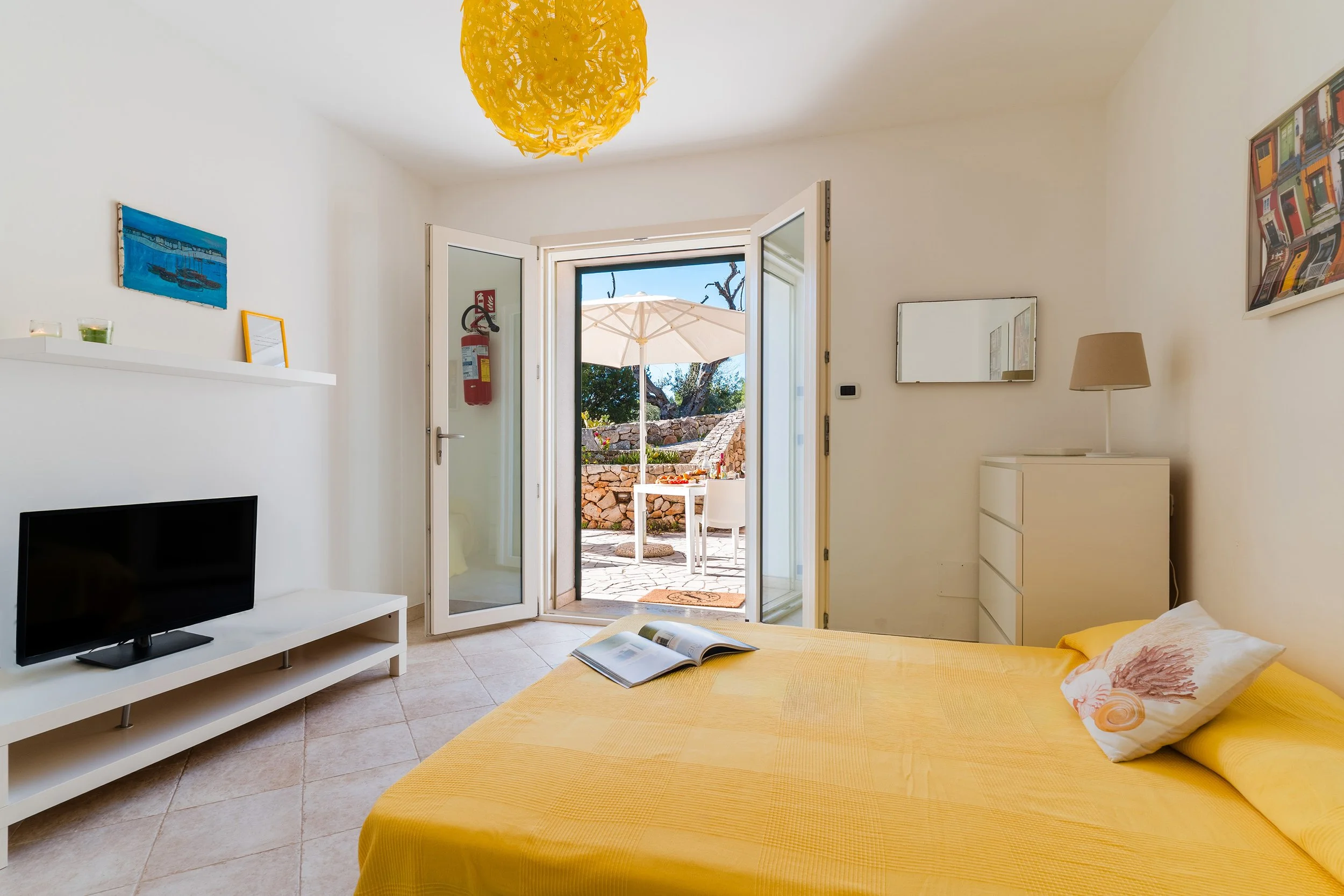 A bedroom with a yellow bedspread and a pillow with a seashell design, an open magazine on the bed, a white TV stand with a flat-screen TV, white shelves with candles and a framed picture, three paintings on the walls, an open door leading to an outd
