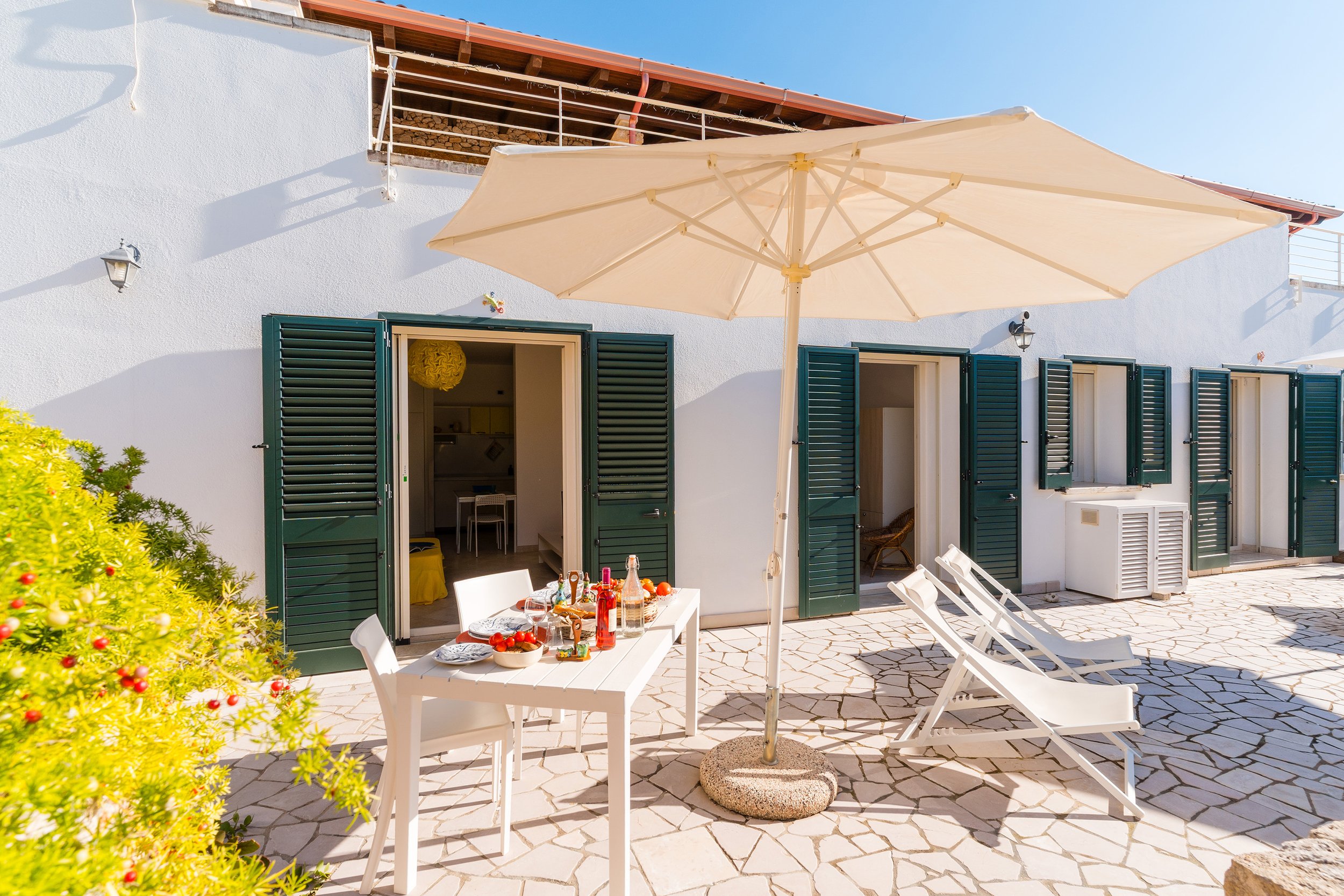 An outdoor patio area with a white table set with drinks and food, surrounded by white chairs, a large beige umbrella, and green plants, in front of a white building with open green shutters and a balcony under a clear blue sky.