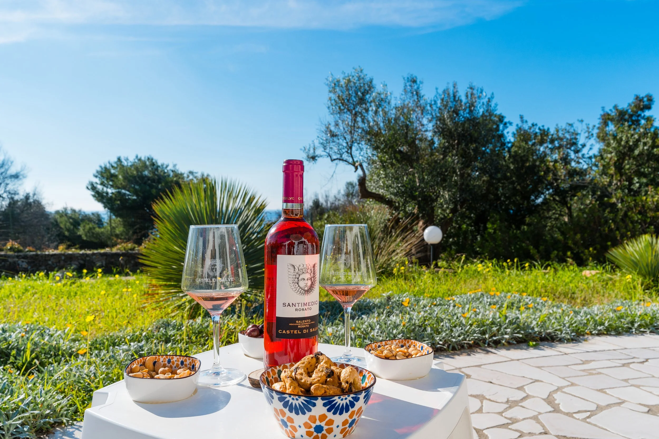 A table set with a bottle of rosé wine, two glasses of wine, bowls of mixed nuts, cookies, and other snacks, outdoors on a garden patio with trees and a blue sky in the background.