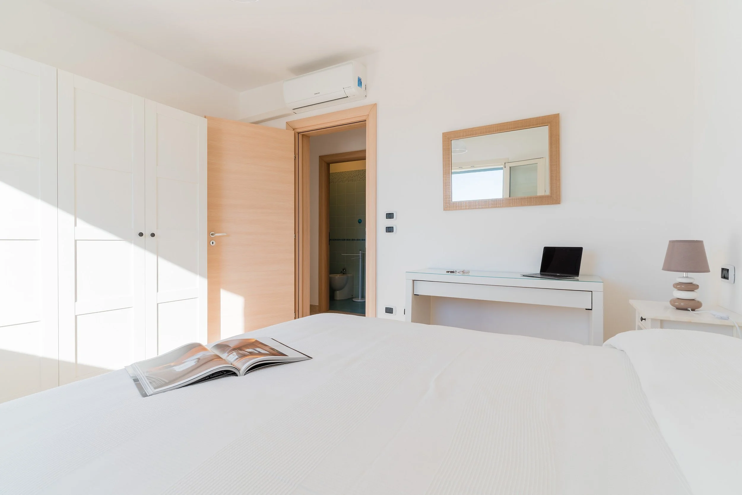 A bedroom with a white bed, an open magazine on it, a white dresser with a lamp, a mirror on the wall, a laptop, a wooden door, white closet, and a small bathroom with a toilet visible through the doorway.