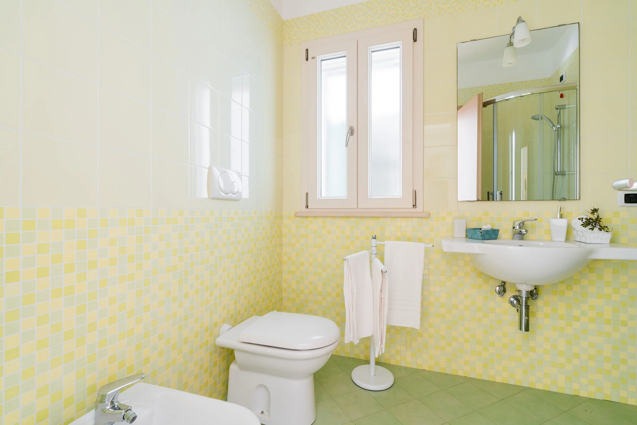 A small bathroom with yellow mosaic tile walls, a white toilet, a small white sink, a wall mirror, a window with blinds, and a shower with a curved glass door.
