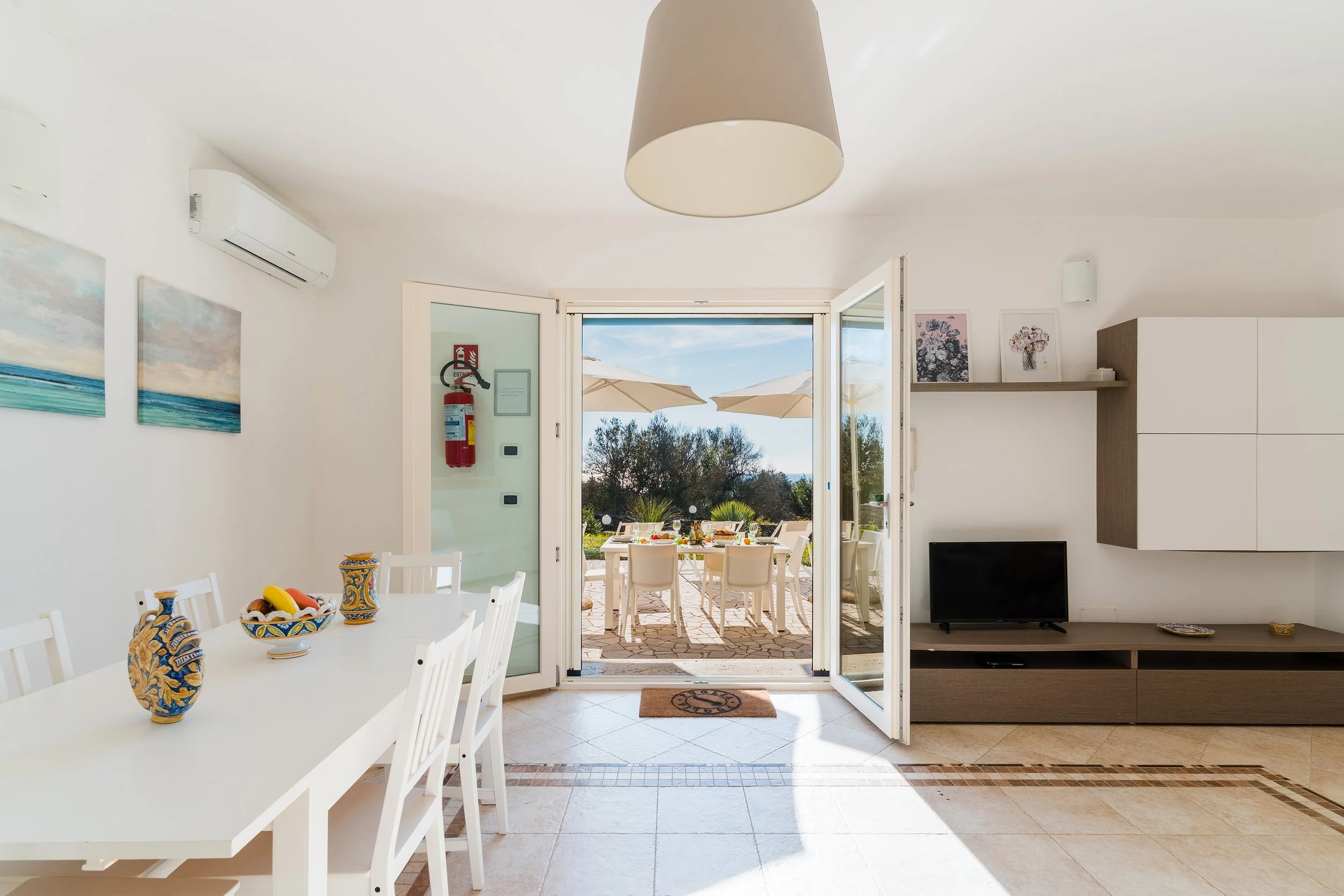 Open door leading to patio with outdoor dining area under umbrellas, view of trees and sky.