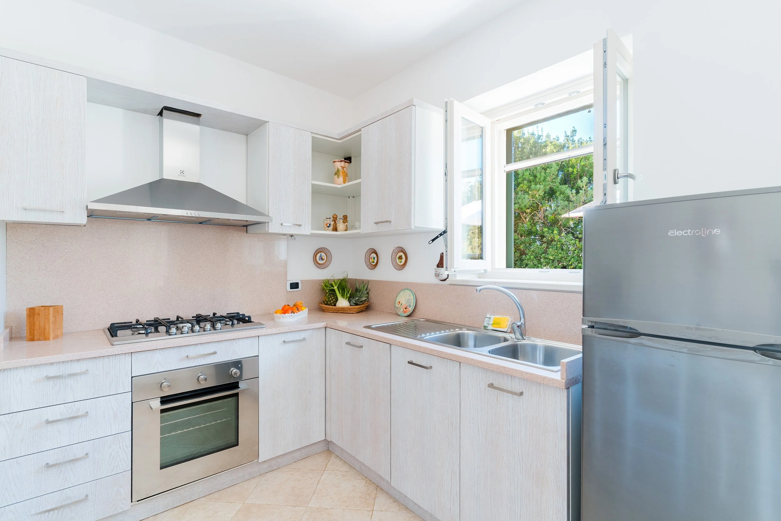 A bright, modern kitchen with white cabinets, a stainless steel oven, and a gas stove. There is a double sink beneath a window that opens outward to a view of green trees. The kitchen has a light pink countertop and decorative plates hanging on the w