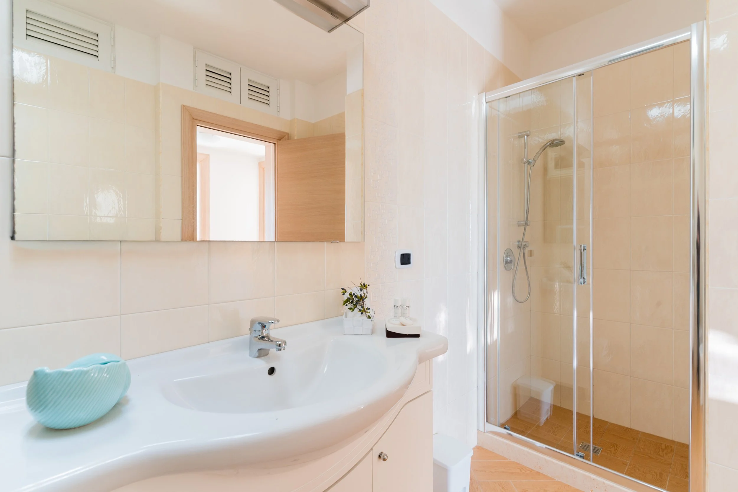 A bright bathroom featuring a white sink with a chrome faucet, a mirror, decorative items including a green vase and toiletries, a shower with glass door and handheld showerhead, and a door with wooden trim.