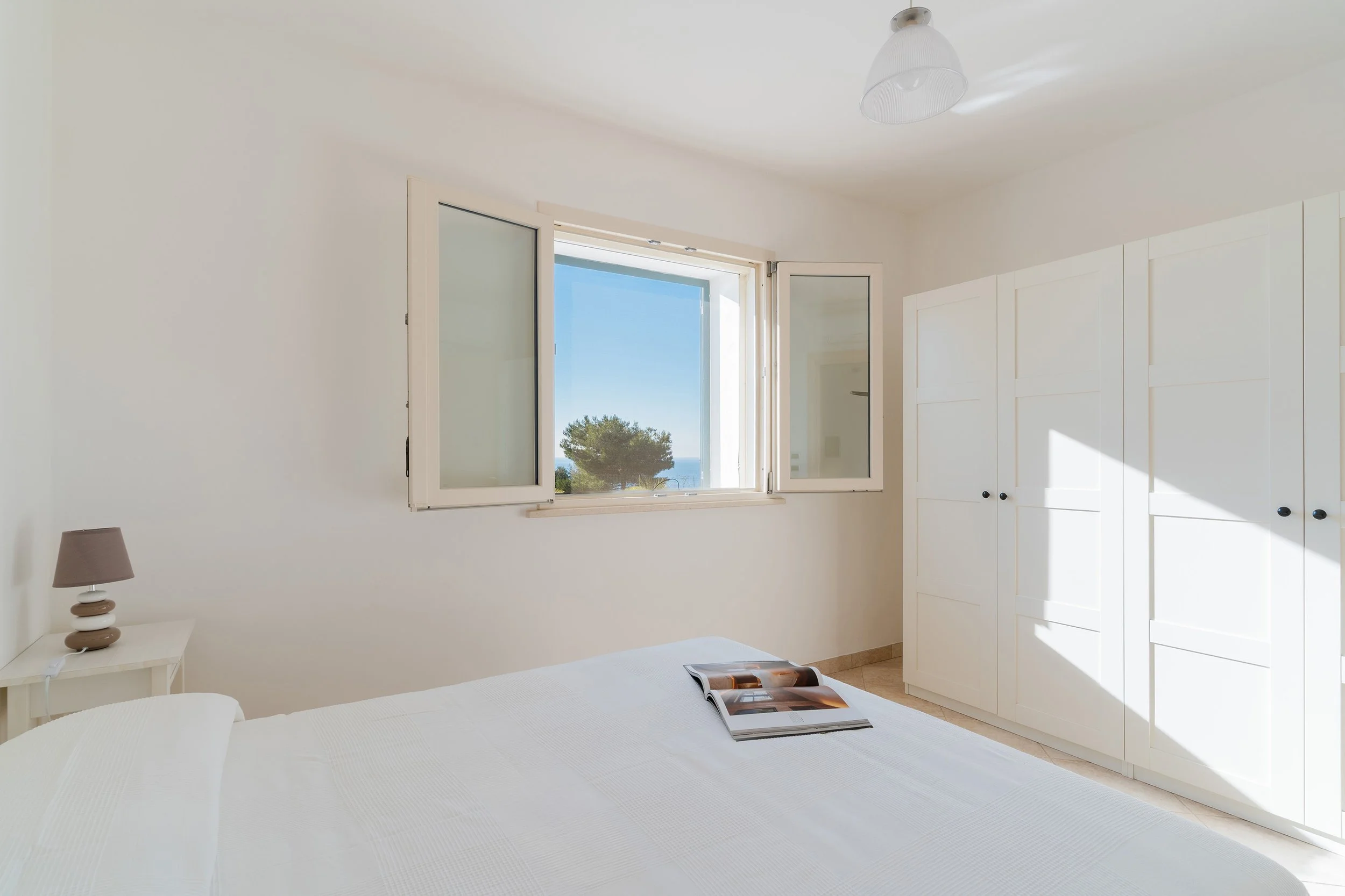 Bright bedroom with white walls, open window showing trees and sky, white wardrobe, small bedside table with a lamp, and an open magazine on the bed.