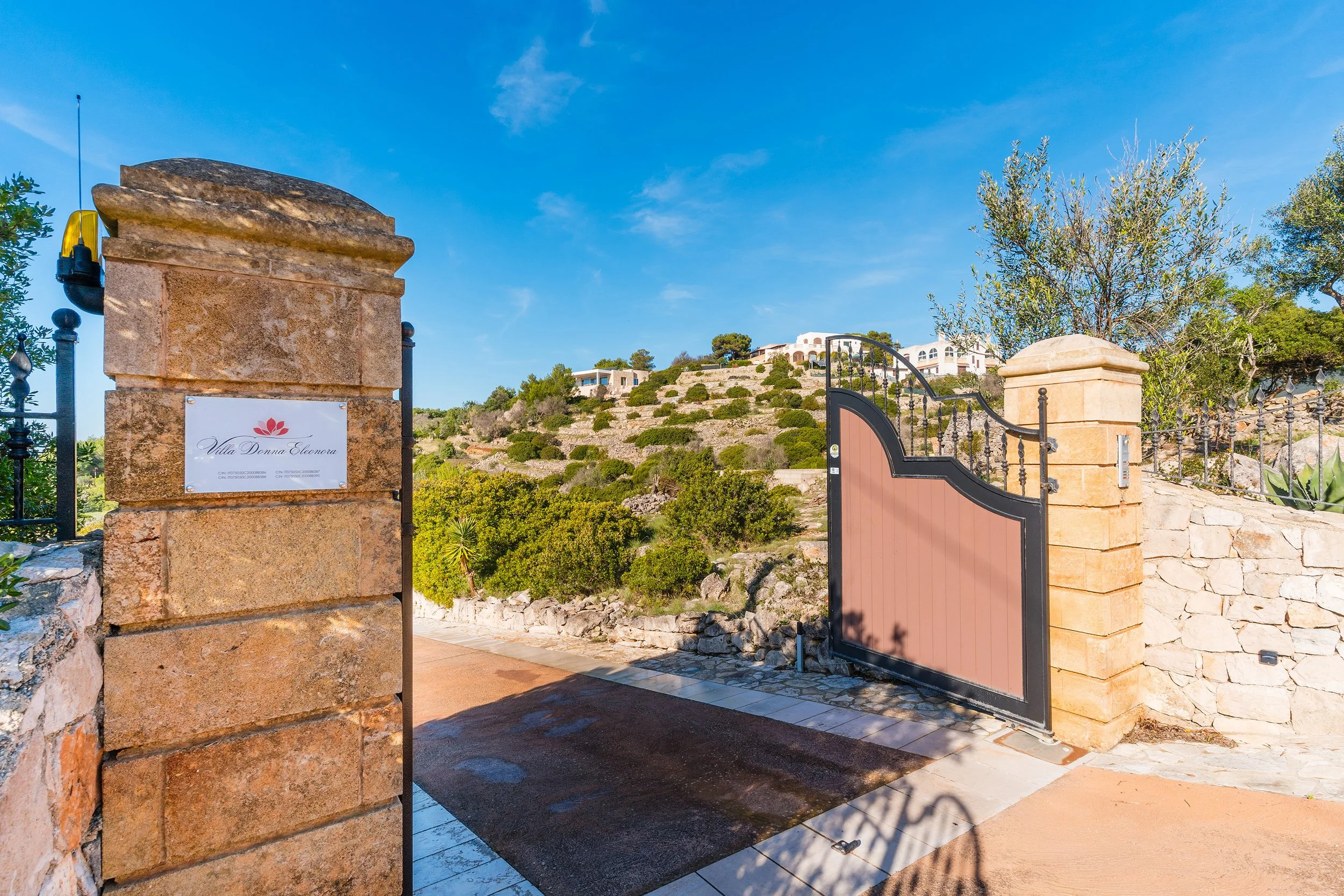 View of a gated entrance with stone pillars and a sign that reads 'Villa Donna Eleonora'; in the background, hillside residential buildings and lush trees under a clear blue sky.