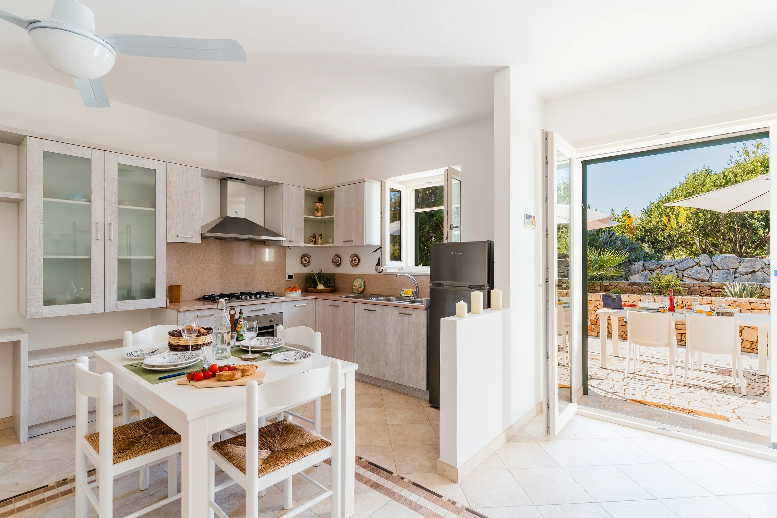 A bright kitchen with white cabinets, a dining table set with plates, glasses, and a tomato-based dish, and an open door leading to an outdoor patio with chairs, an umbrella, green trees, and a stone wall.