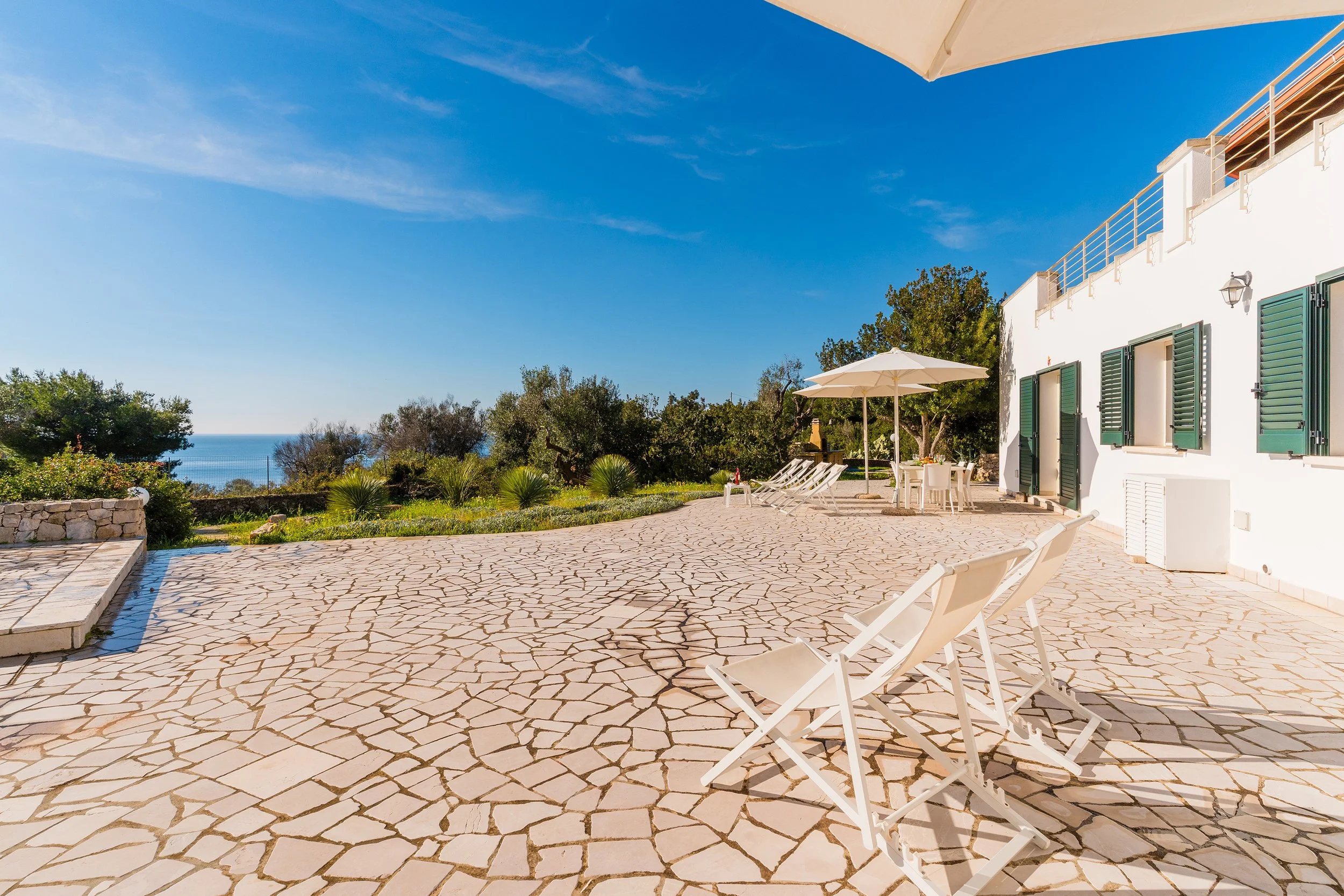 A sunny outdoor patio with white chairs, a large umbrella, green shutters on a white house, and a scenic view of trees and the ocean in the distance.