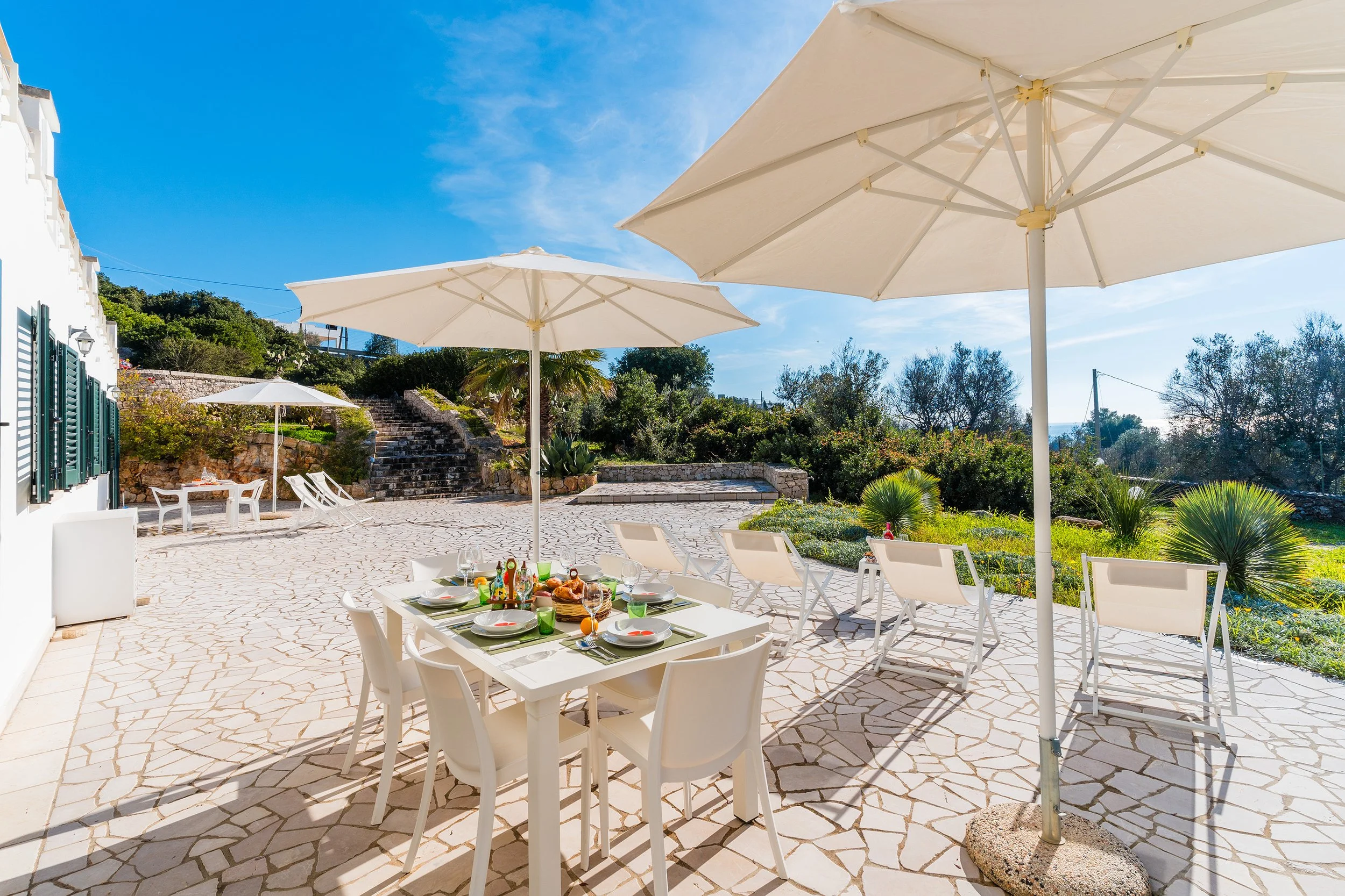 Outdoor dining area with a table set for a meal and white chairs under large white umbrellas, overlooking a garden and stone steps leading up a hill, with a blue sky overhead.