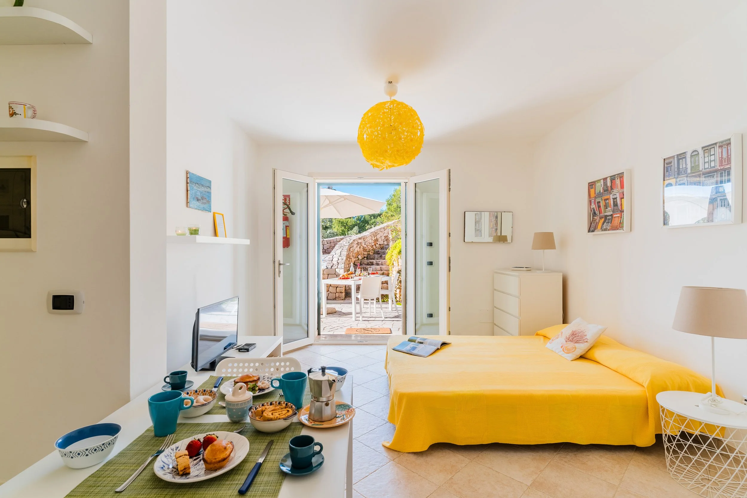 Bright living room with open doors leading to an outdoor patio, yellow sofa, white walls, modern artwork, and a dining table set with breakfast food and drinks.