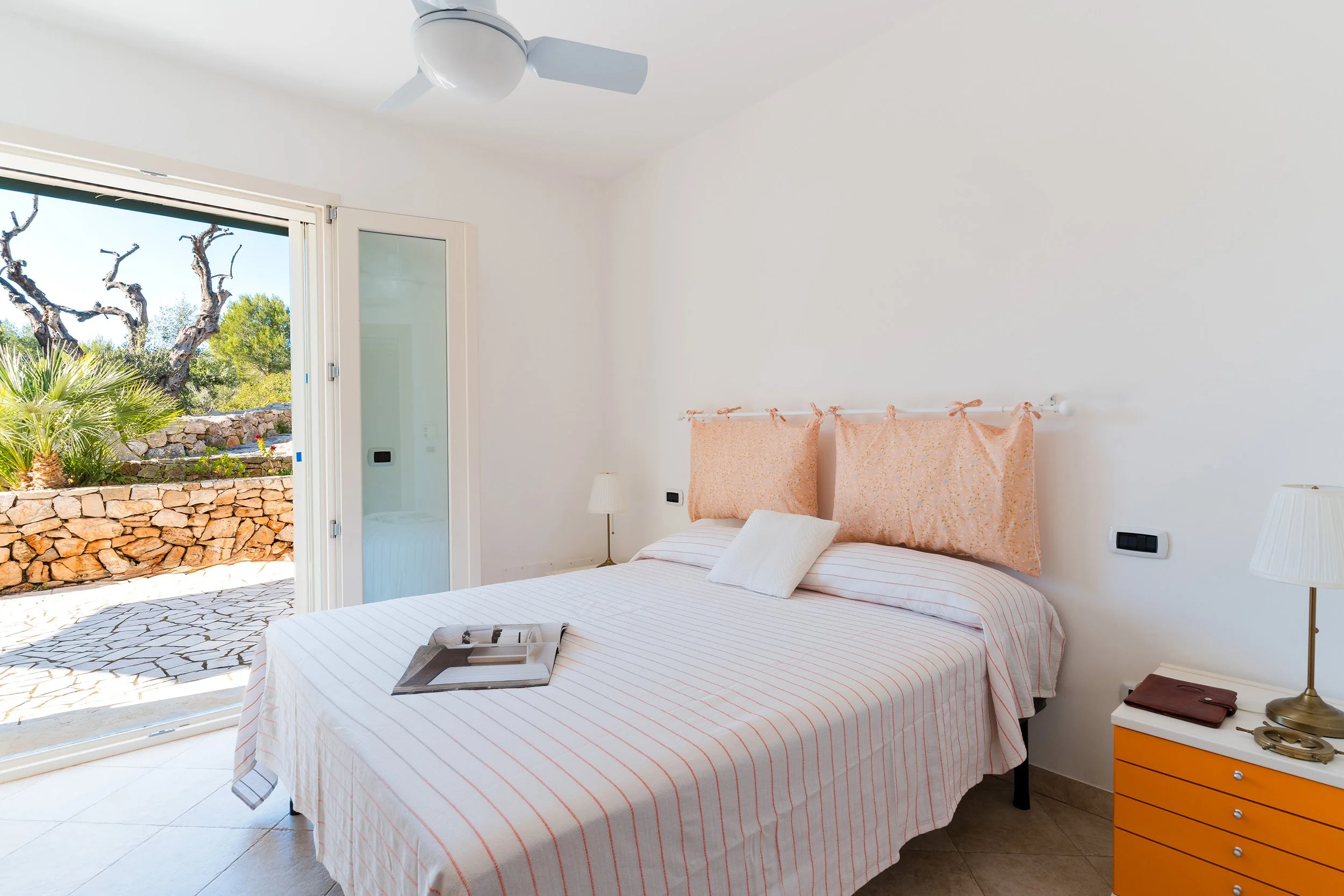 A bedroom with a bed near a sliding glass door, that opens to an outdoor area with a stone wall, plants, and a tree. The bed has a striped bedspread and a pink headboard. There are two nightstands with lamps on each side of the bed, and a yellow dres