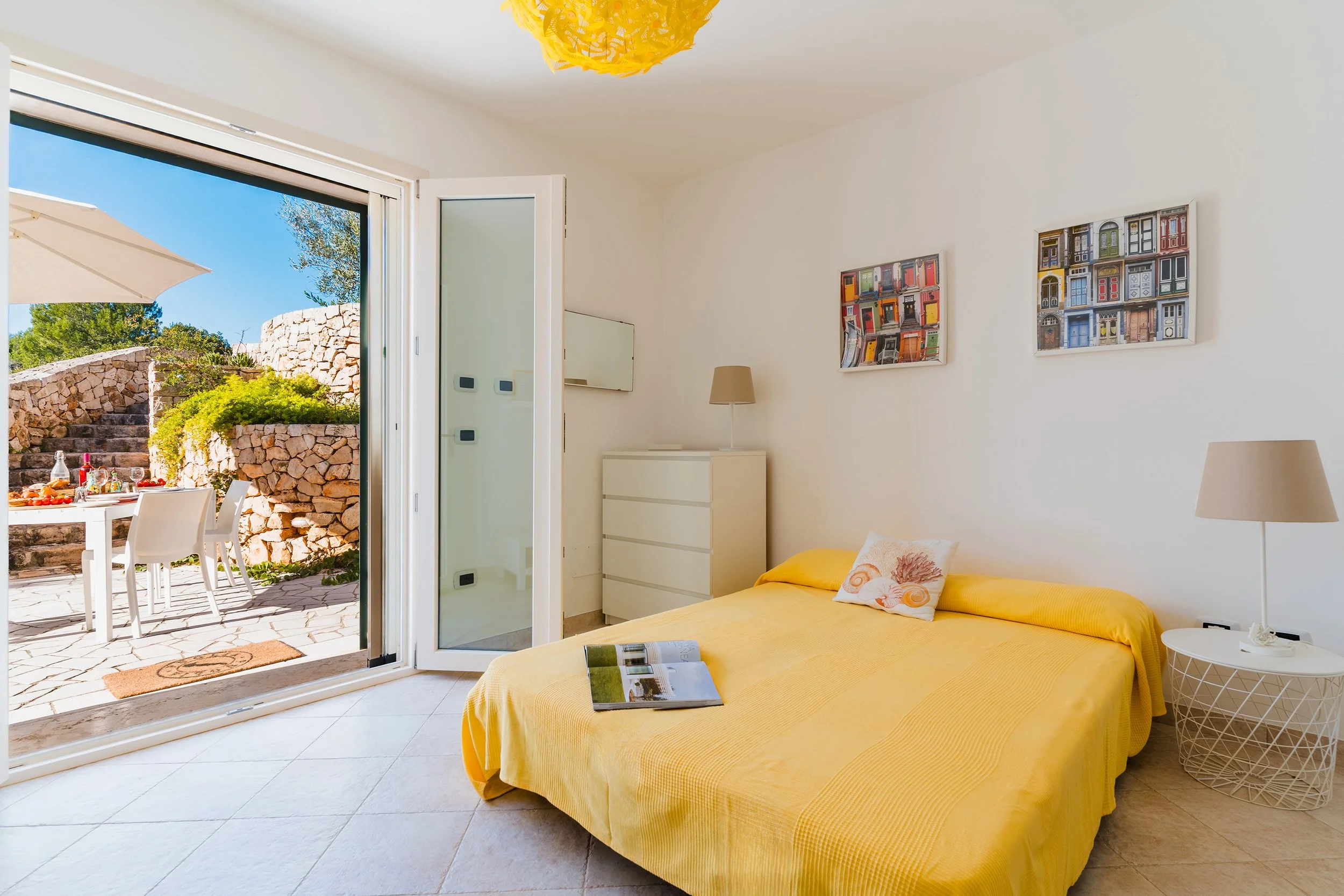 A bedroom with a large glass door open to an outdoor patio with a stone wall, table, and chairs. Inside, there is a bed with yellow bedding, a white dresser, a lamp, and framed artwork on the wall.