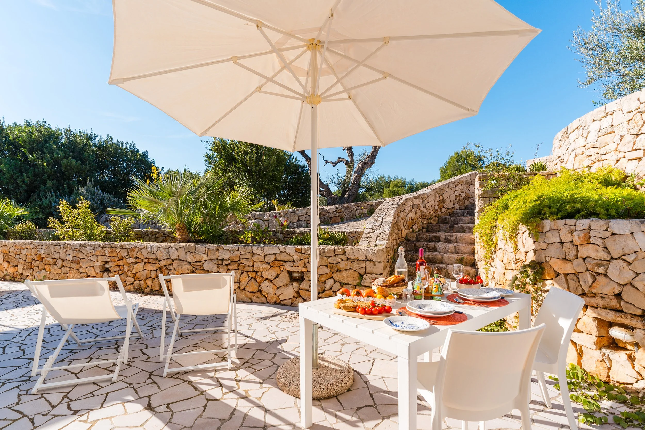 Outdoor dining area with a white table set with food and drinks, surrounded by white chairs, under a large beige umbrella, with a stone wall, green plants, and trees in the background on a sunny day.