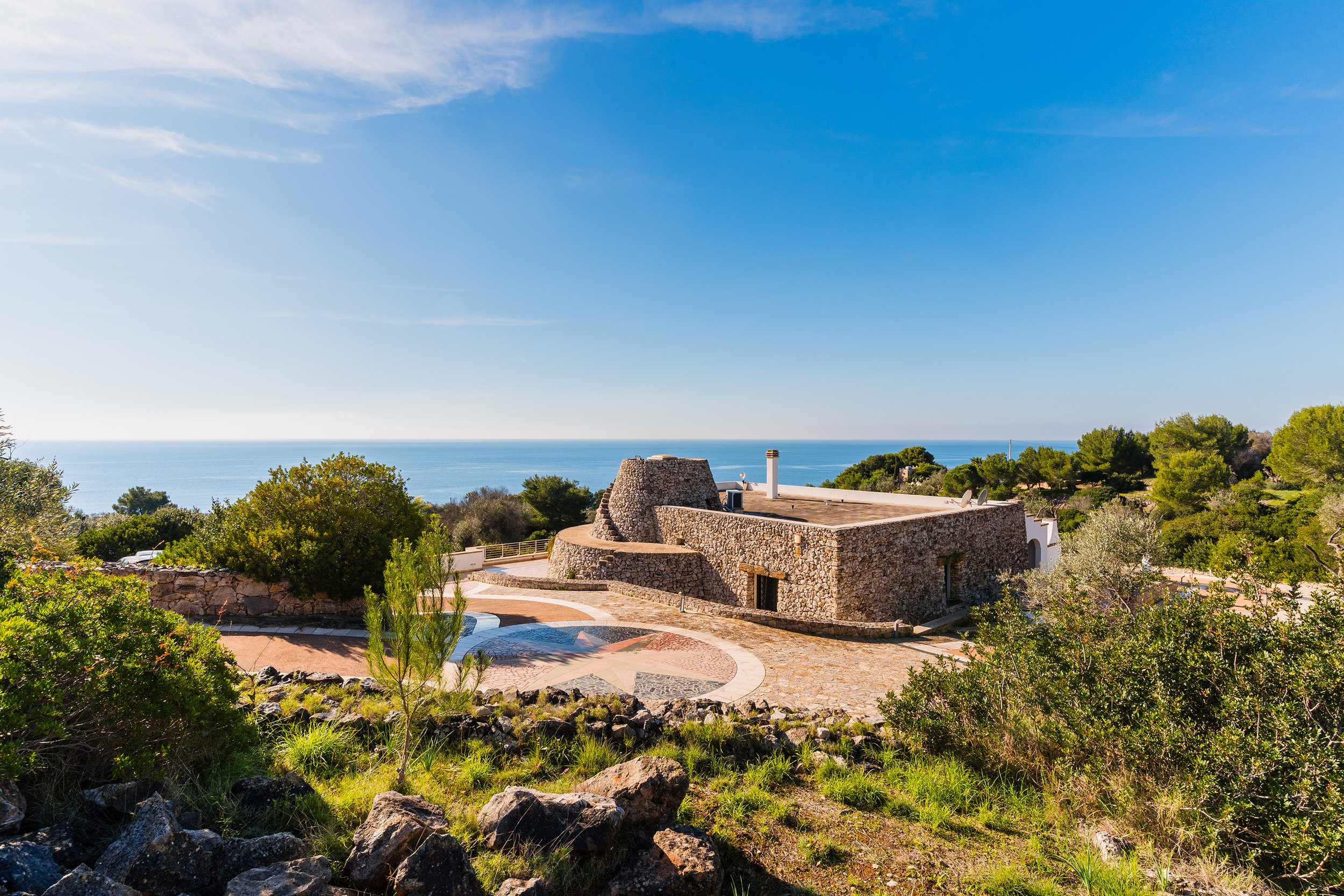 A scenic view of a stone house with a chimney among green trees under a clear blue sky, overlooking a large body of water in the distance.