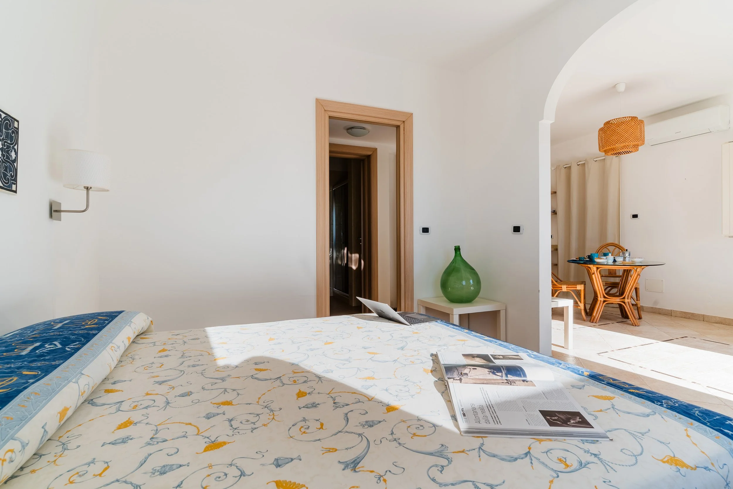 A bedroom with a bed covered in white and yellow patterned bedspread, magazine and newspaper on it, a green glass vase on a white side table, open doorway leading to a hallway, and visible dining area with wooden table and chairs, orange pendant ligh
