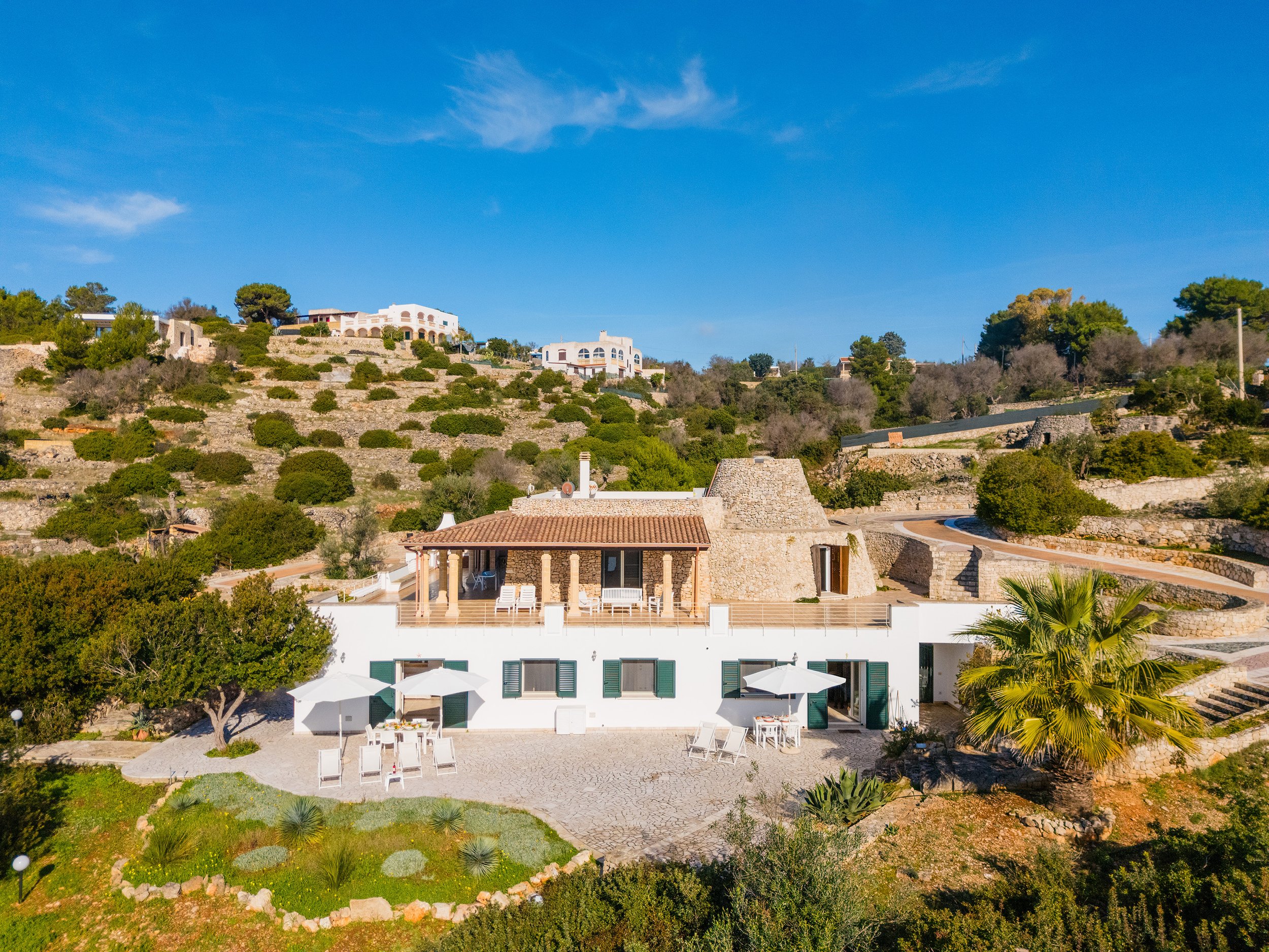 A white house with green shutters and a tan tiled roof, surrounded by a garden with outdoor dining tables and chairs, a palm tree, and other plants, with a hillside of houses and trees in the background under a clear blue sky.