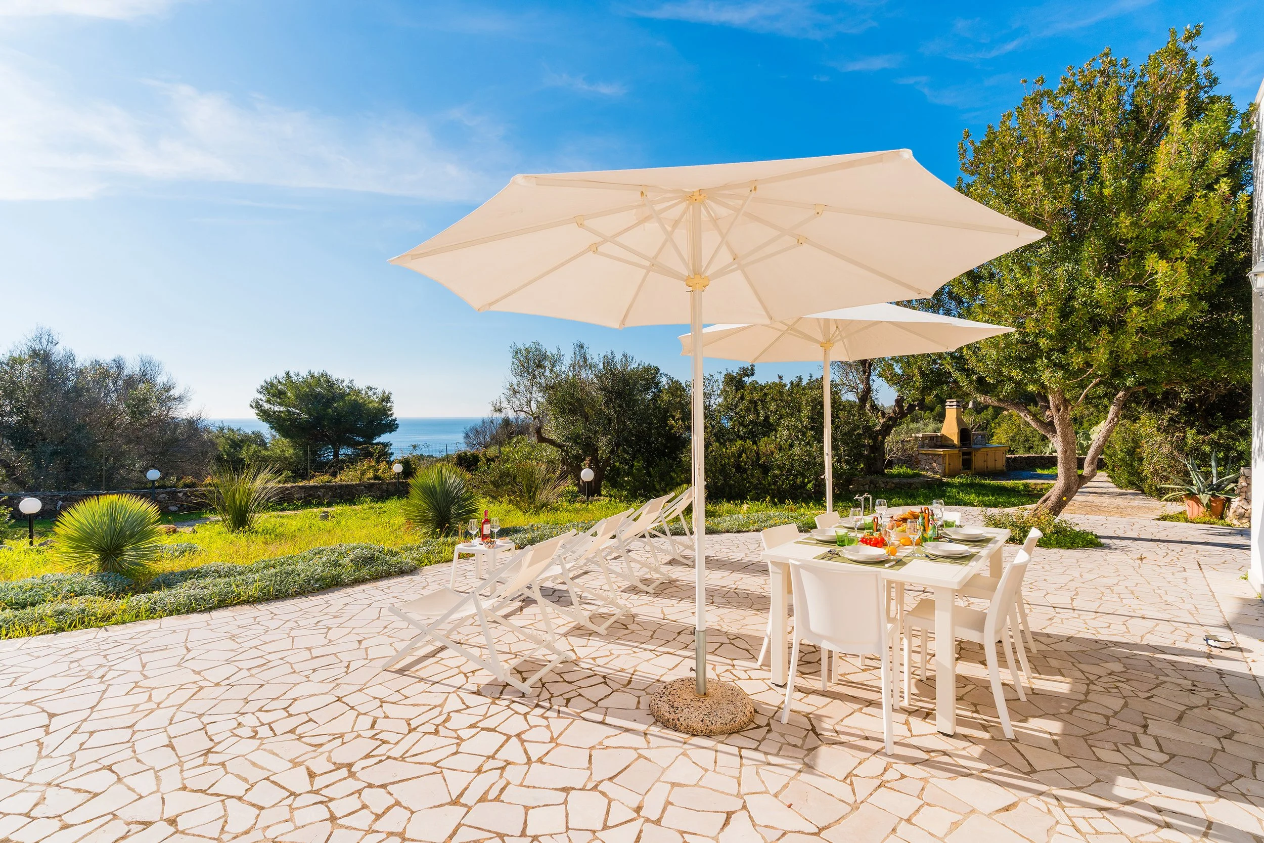 Outdoor patio set with white table, chairs, and large white umbrellas, surrounded by greenery and trees under a bright blue sky.