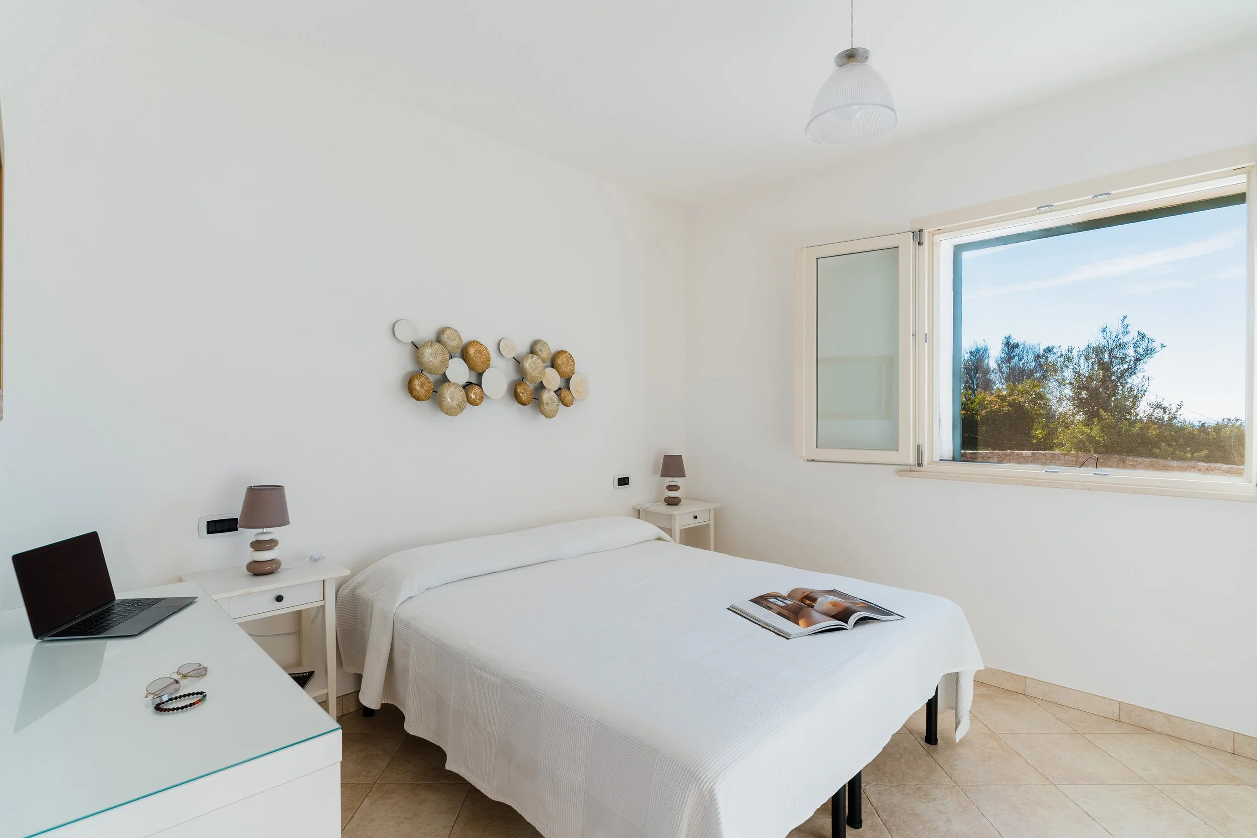 A bright bedroom with a white bed, nightstand, and lamps. An open window shows a view of trees and blue sky.