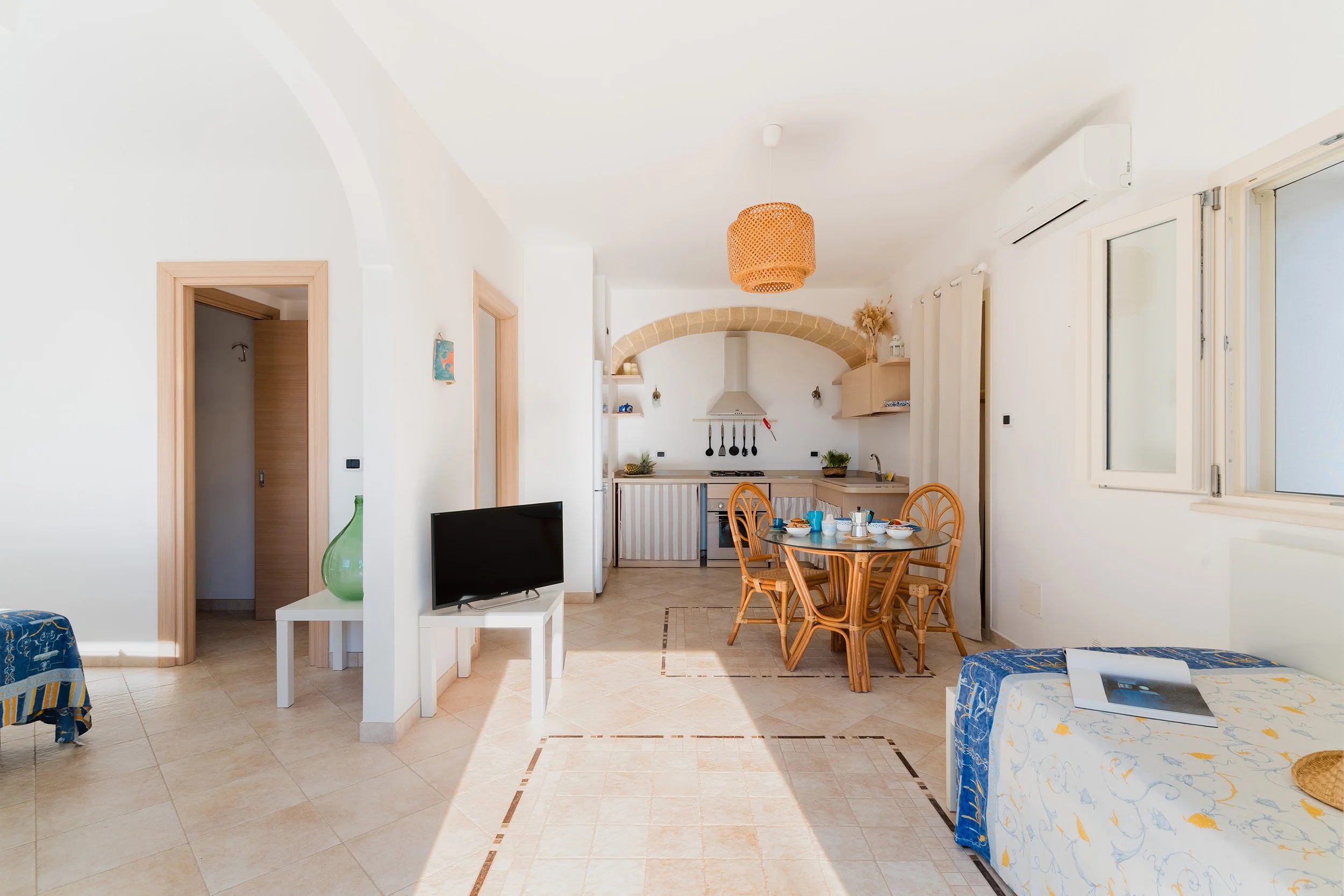 Open living room with dining area, kitchen in the background, and a window on the right. The room has white walls, tiled floor, and wooden accents. There's a round dining table with wooden chairs, a small flat-screen TV, and a ceiling light with a wo