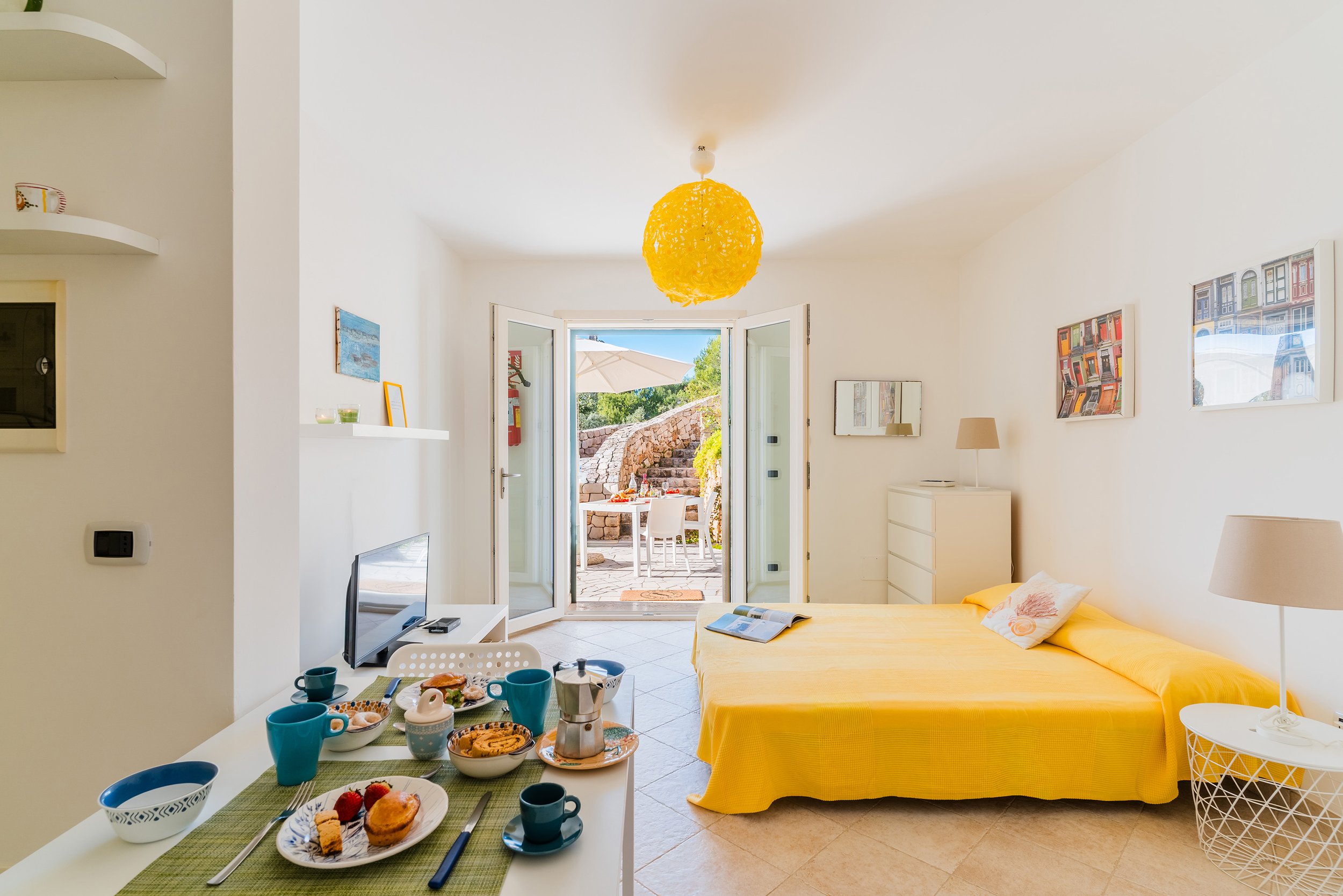 An open-concept living room and dining area with a bright yellow bed, a breakfast table set with food and mugs, and an outdoor patio with seating and an umbrella visible through open double doors.
