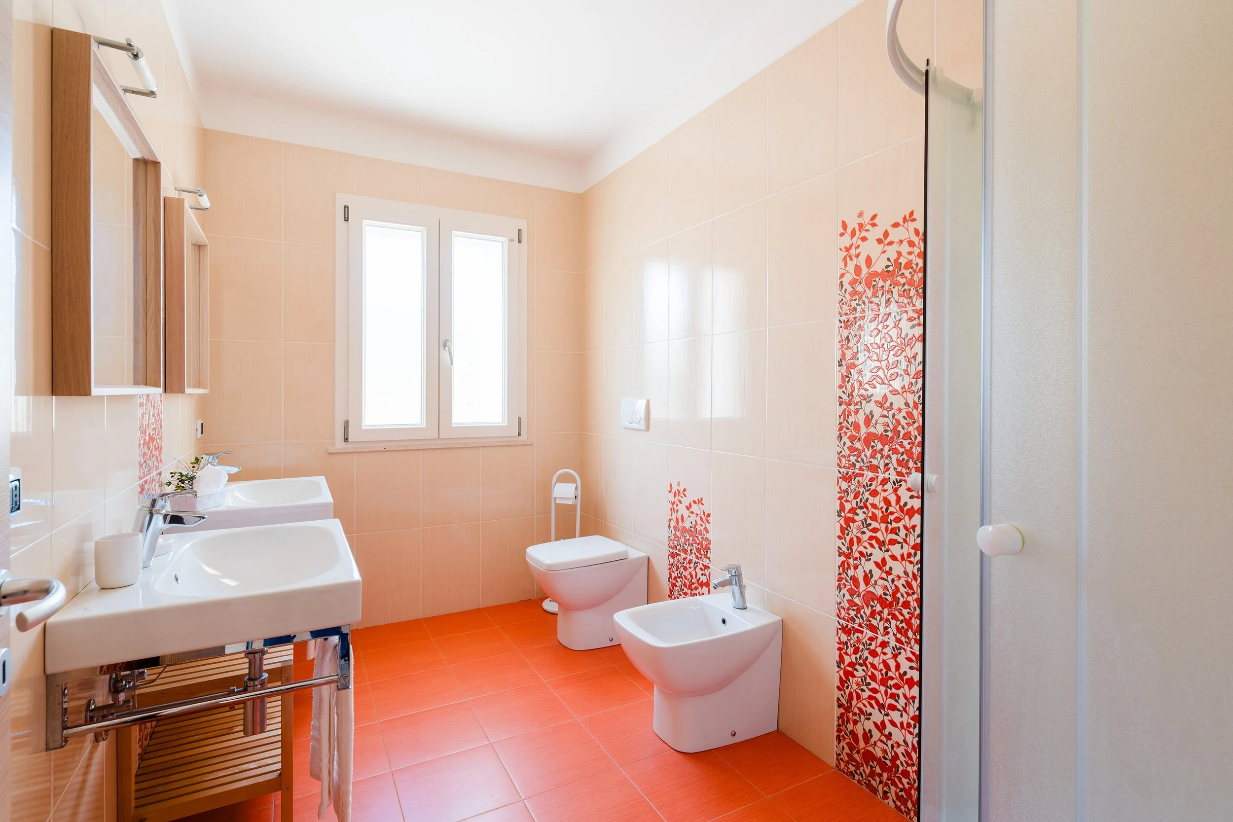 Bright bathroom with peach-colored tiled walls, orange floor tiles, a white sink with a chrome rack, a window, a toilet, and a bidet. There is a decorative red and white floral tile pattern behind the shower.