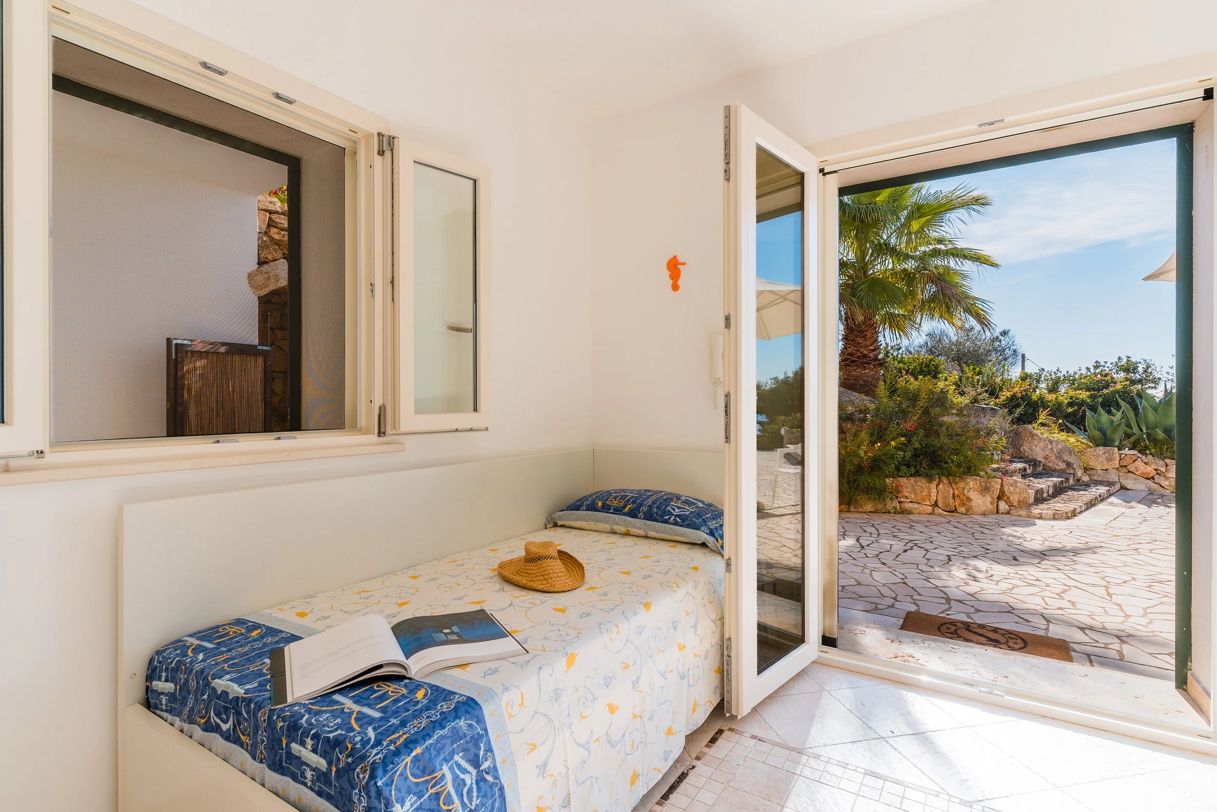Open white-framed glass door leading to a patio with stone paving, outdoor plants including a palm tree, and a partly cloudy sky.