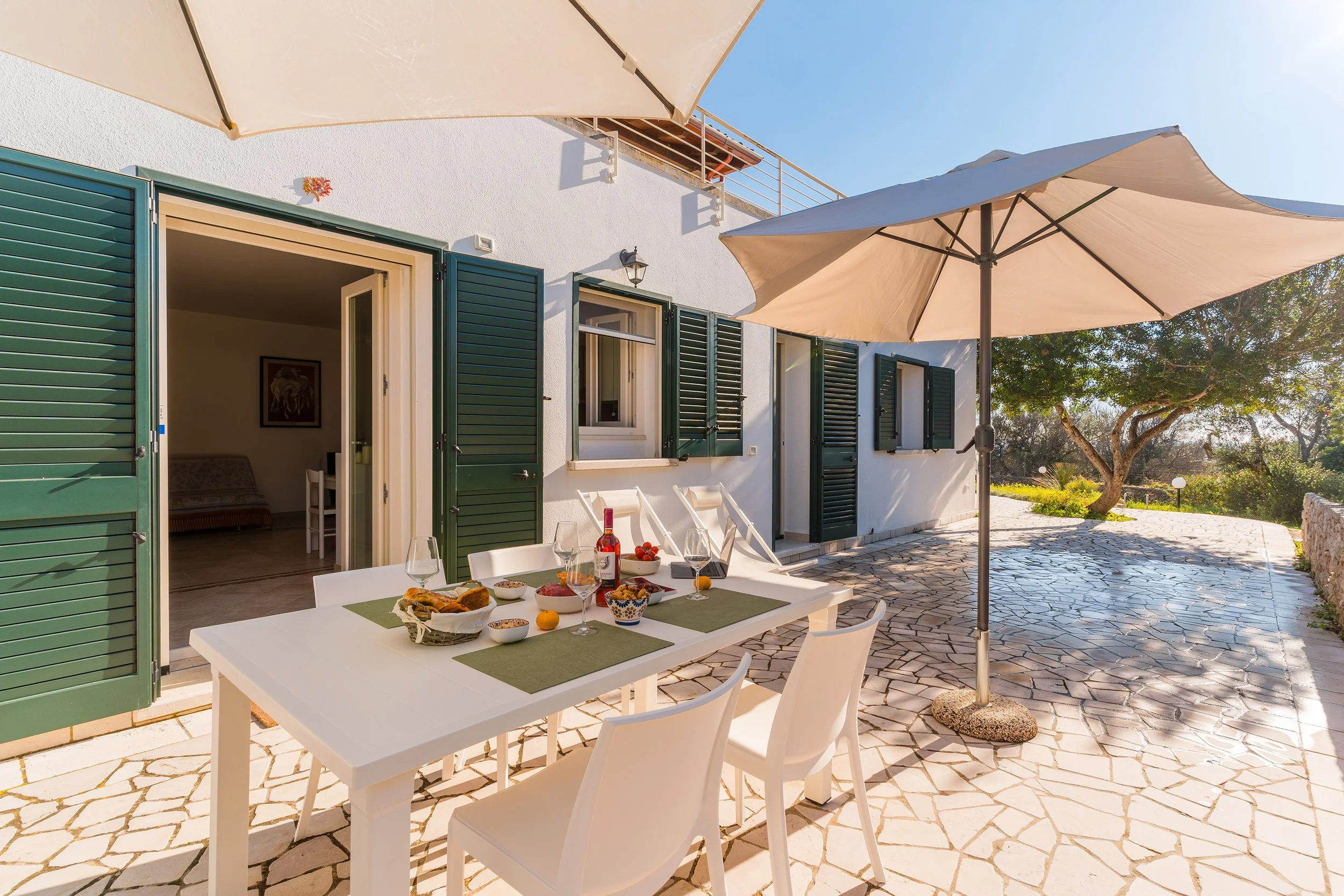 Outdoor patio dining area with a table set for breakfast, featuring wine glasses, a bottle of wine, bowls of fruit and pastries, and a small lemon, under white umbrellas. The background shows a white building with green shutters, open doors, and a co