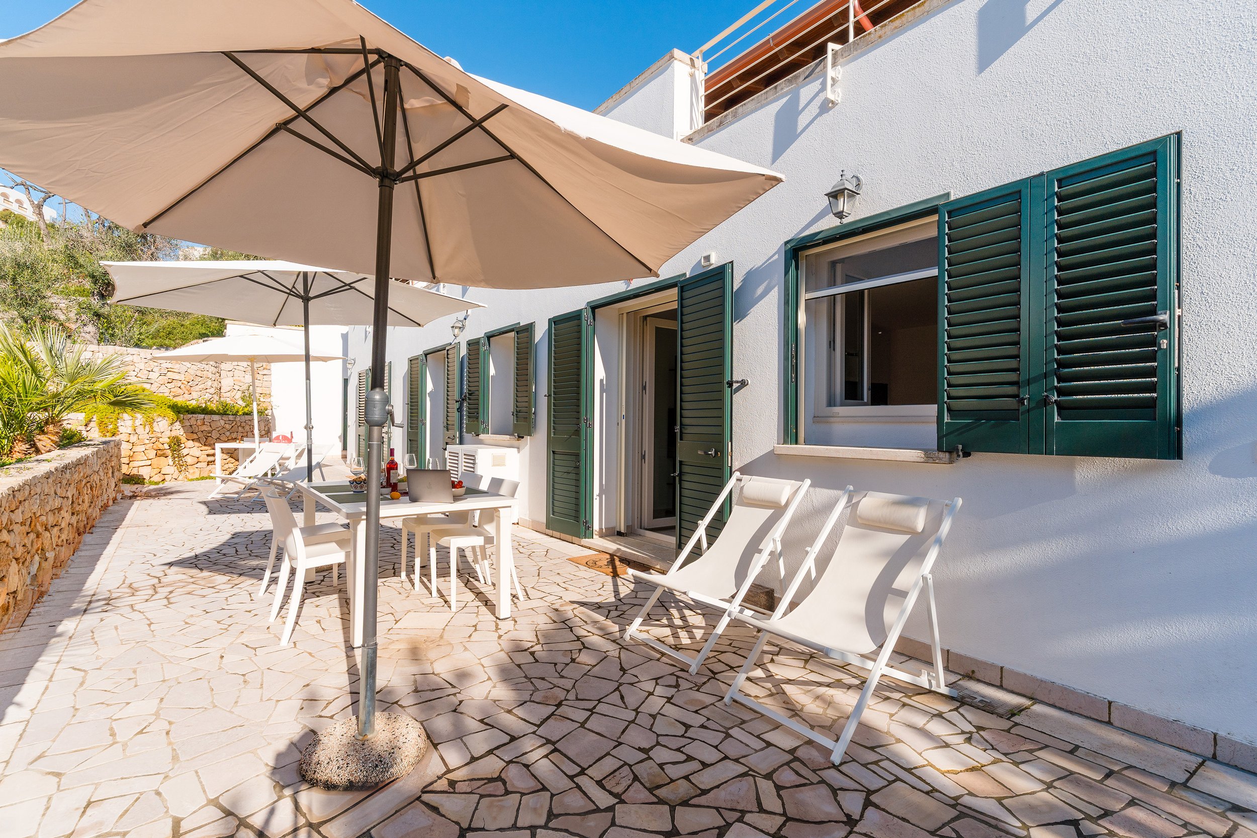 Outdoor patio area with white chairs, umbrellas, and a stone pathway outside a white building with green shutters and a balcony.