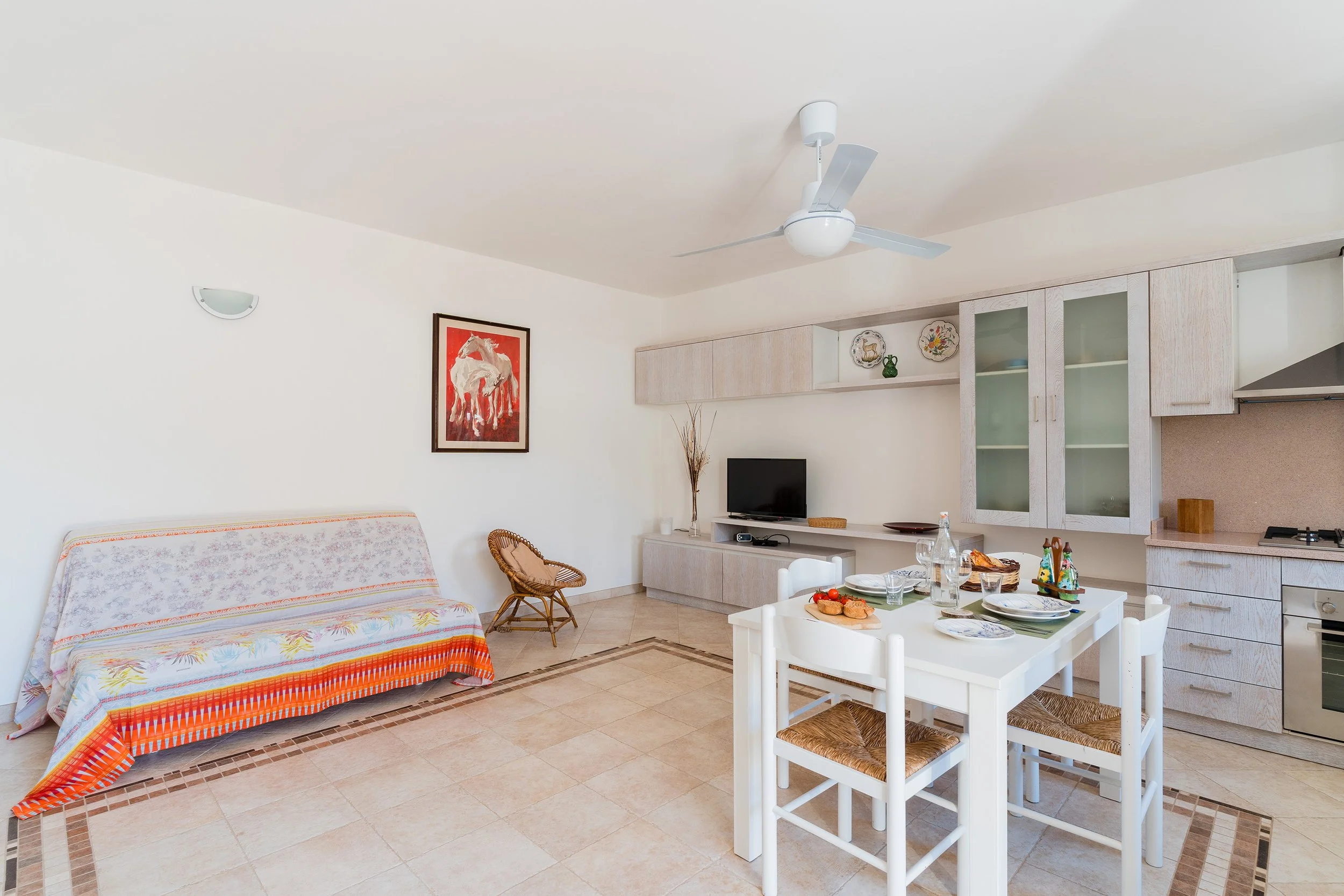 A living room with a white ceiling fan, a framed art piece of horses on the wall, a floral patterned sofa with a colorful cover, a wicker chair, a small flat-screen TV on a white cabinet, a table with food, drinks, and colorful decorations, and a kit