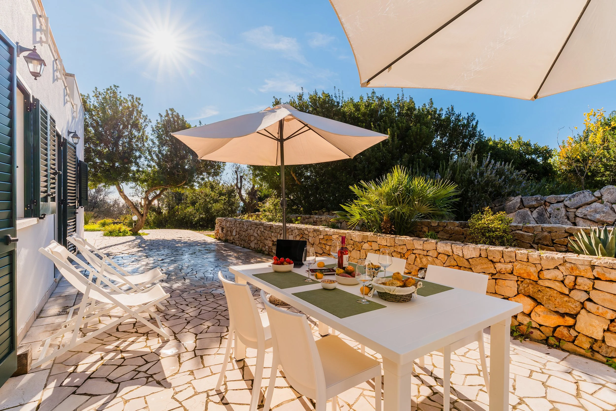 An outdoor dining area on a stone patio with a white table set with food and drinks, surrounded by white chairs, large umbrellas providing shade, a stone wall, lush greenery, and sunny blue skies.