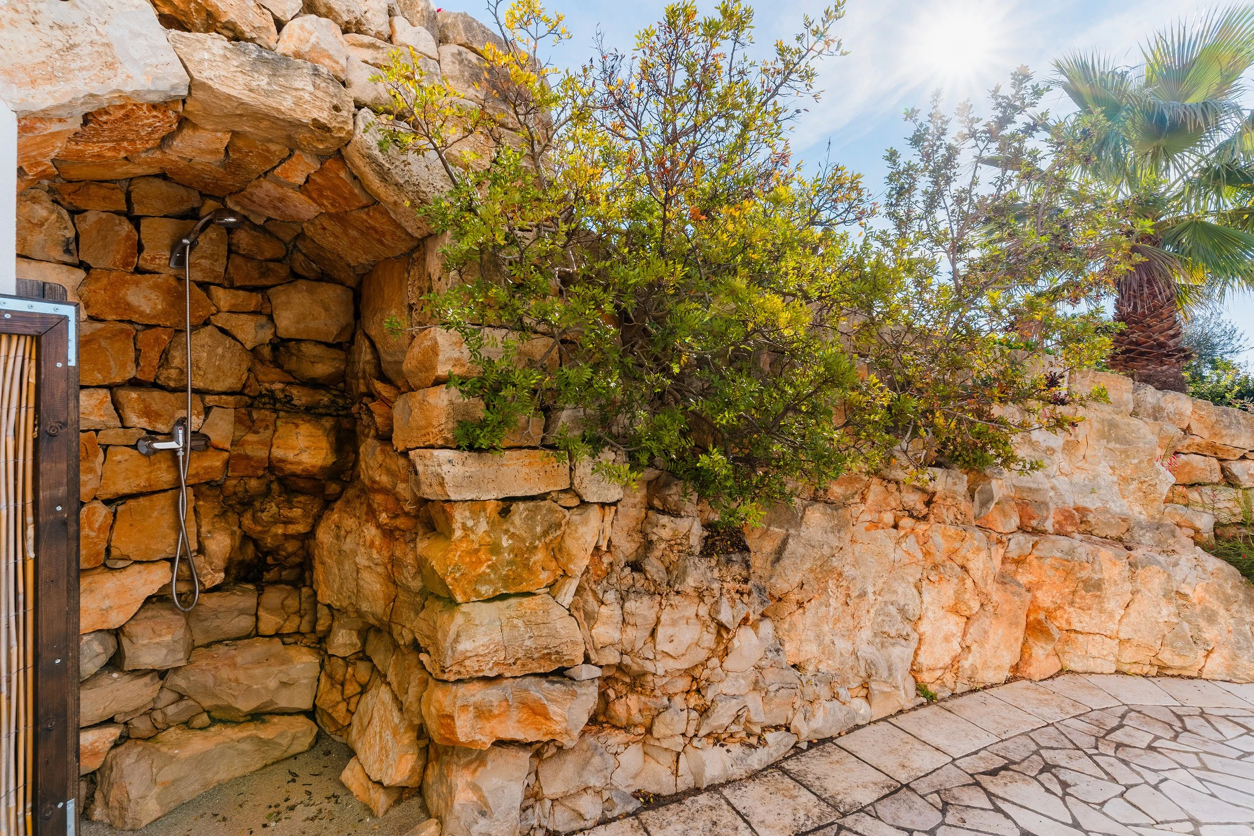 Outdoor shower area built into a stone wall with trees and bright sunlight in the background.