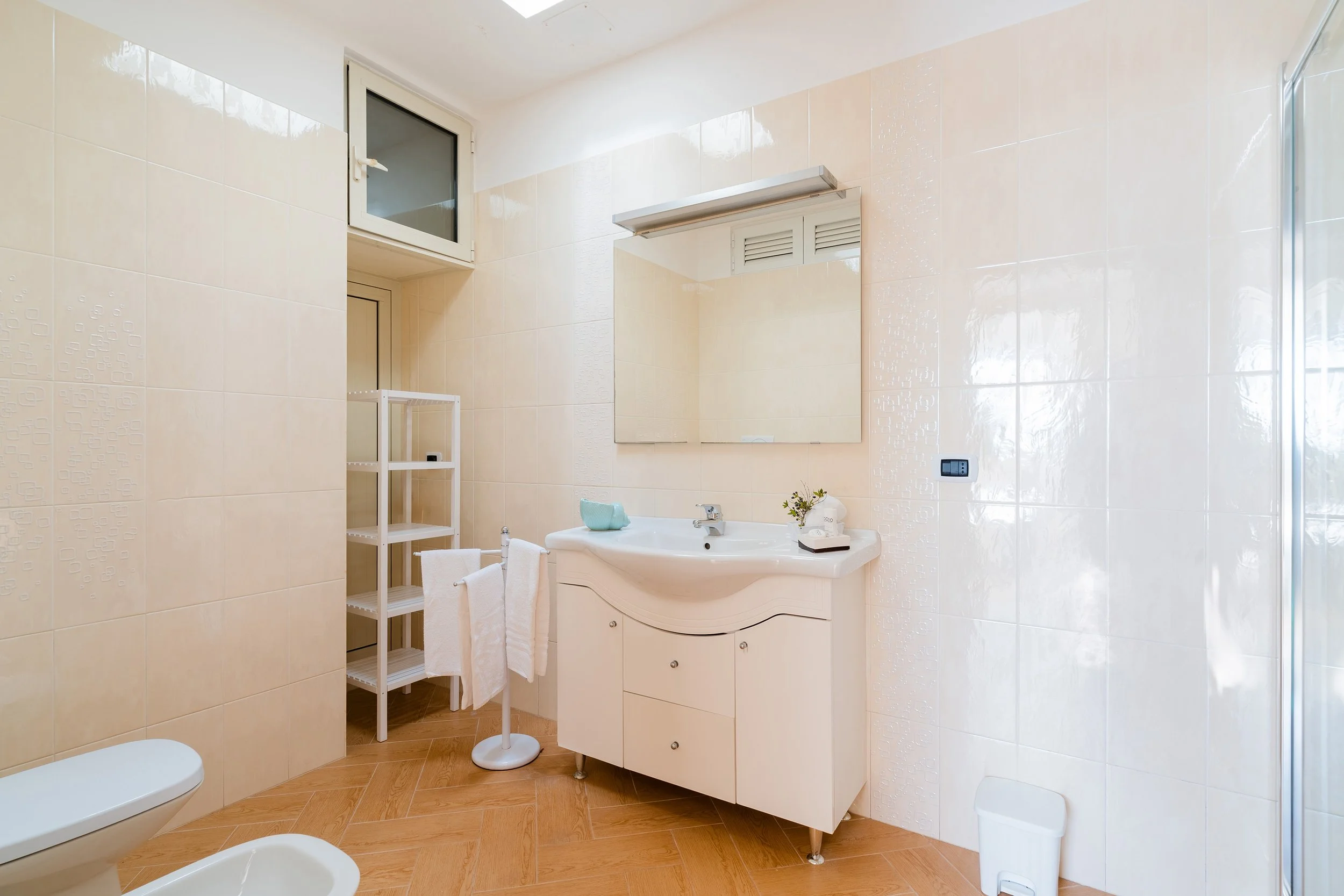 Clean bathroom with beige tiled walls, a white sink with a cabinet, a mirror, a small shelving unit, a towel holder, and a window.