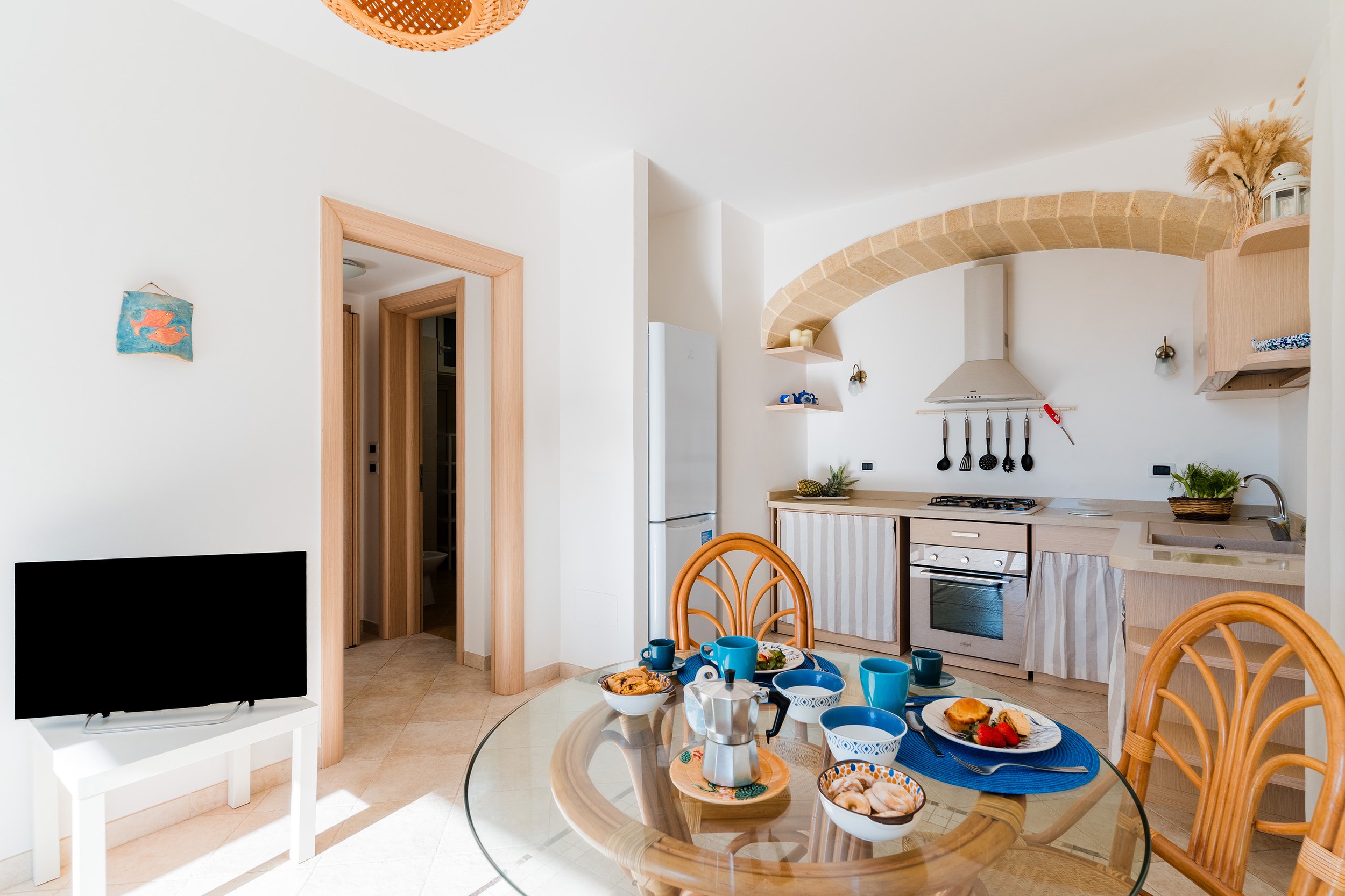 A kitchen and dining area with a round glass table set for breakfast, featuring blue dishes and pastries. The kitchen has light wood cabinets, a white range hood, and black cookware hooks. There are orange wooden chairs, a white TV stand with a black TV, and a pineapple on the counter. The room has white walls, a white refrigerator, and a decorative arch with tan bricks. A doorway and some wall shelves are also visible.
