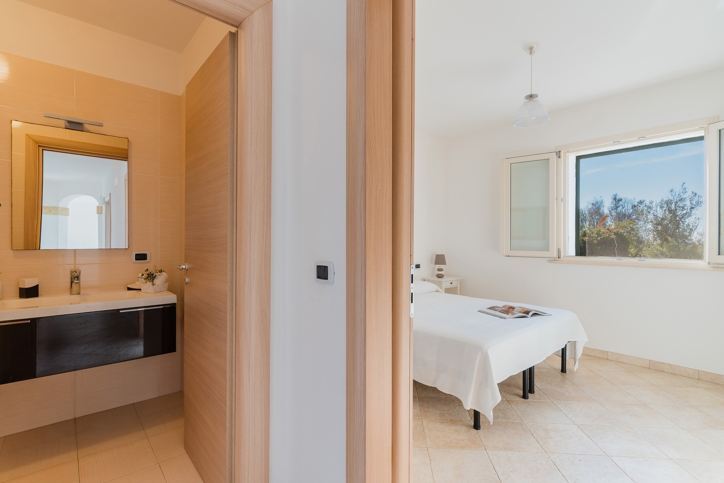 A bedroom with a large window showing trees outside, a white bed with a magazine on top, and a nightstand with a lamp. The room is illuminated by natural light and has a minimalist design with white walls and beige tile flooring.