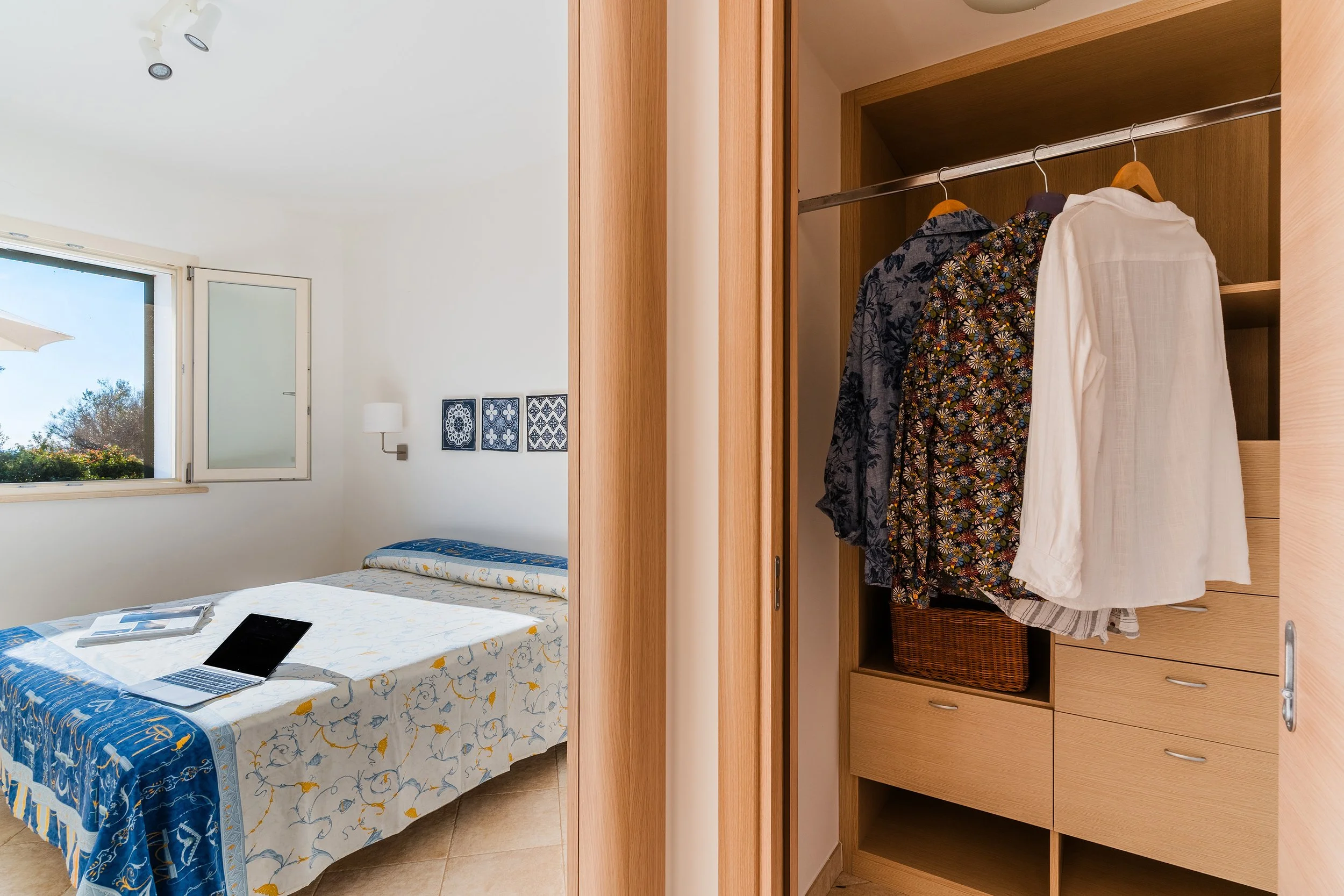 Interior of a bedroom showing a bed with patterned bedspread, an open window with a view of blue sky and trees, framed wall art, a wall-mounted lamp, and an open wooden wardrobe with hanging clothes and a basket inside.