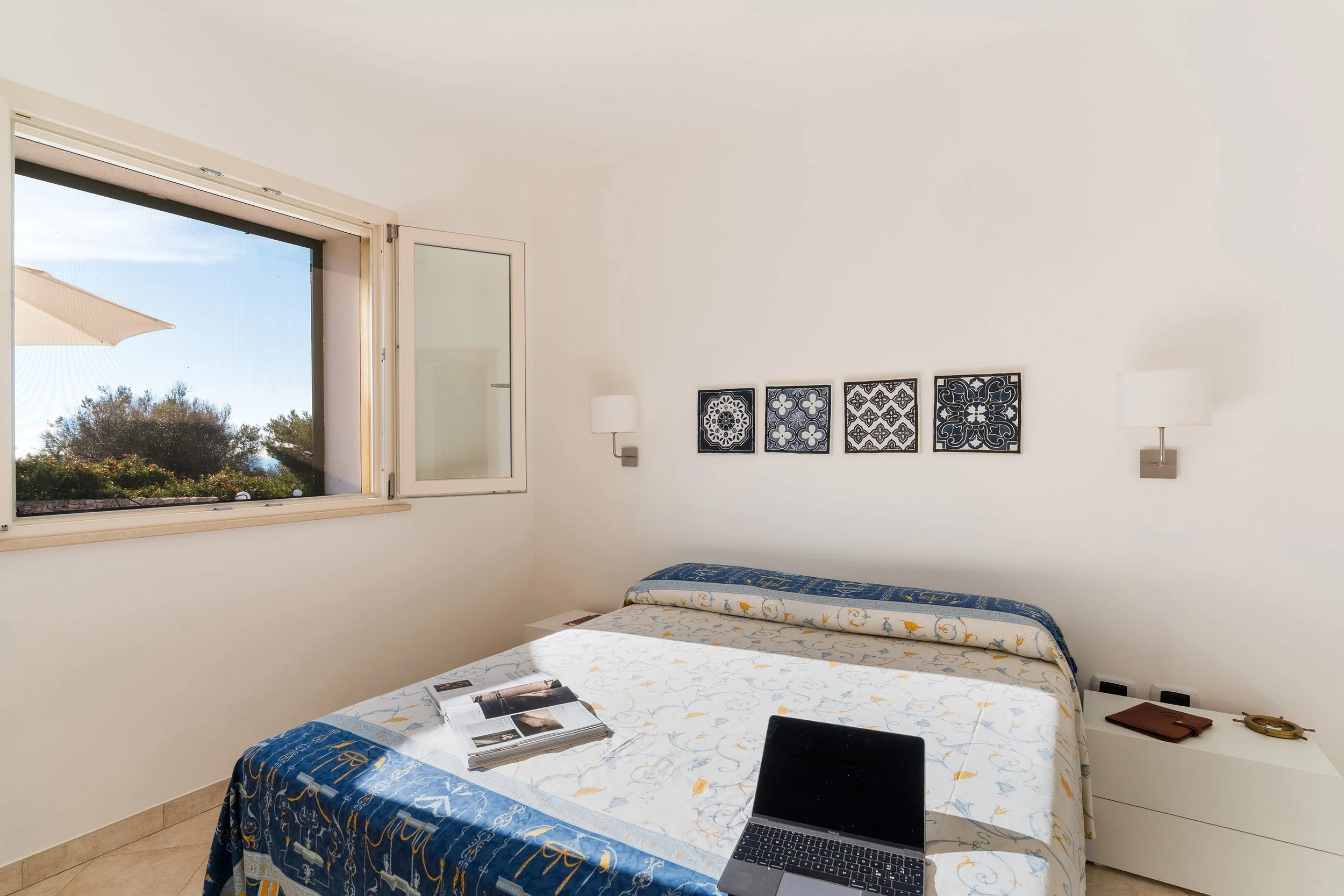 A bedroom scene with a window showing a sunny outdoor view with trees and a beige umbrella. Inside, there is a bed with a blue and white patterned bedspread, an open magazine, and a laptop on top. The wall has four decorative black and white framed t
