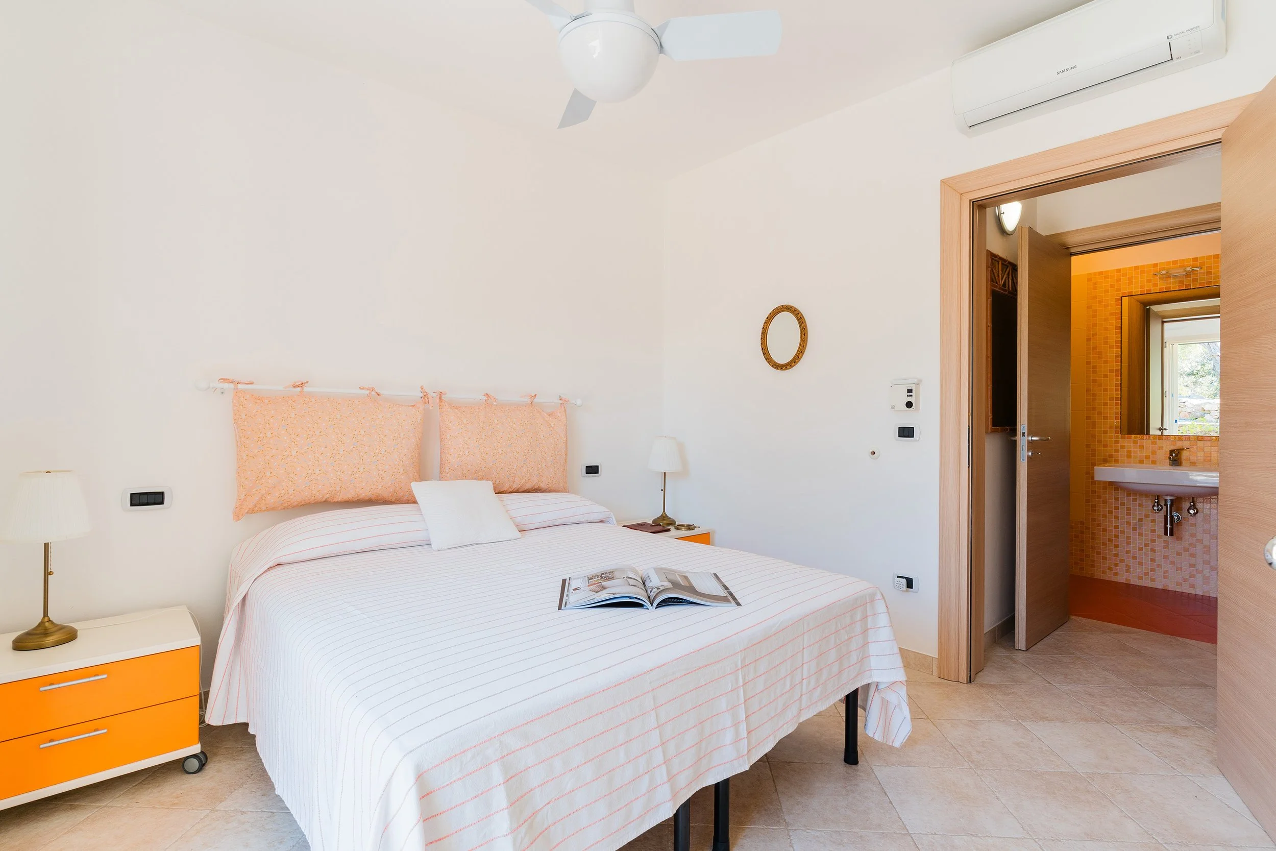 A bedroom with a white bed, orange nightstand, and matching headboard, with a ceiling fan and an air conditioner. There is a small lamp on the nightstand and an open magazine on the bed. The room has wooden doorframes and a doorway leading to a bathr