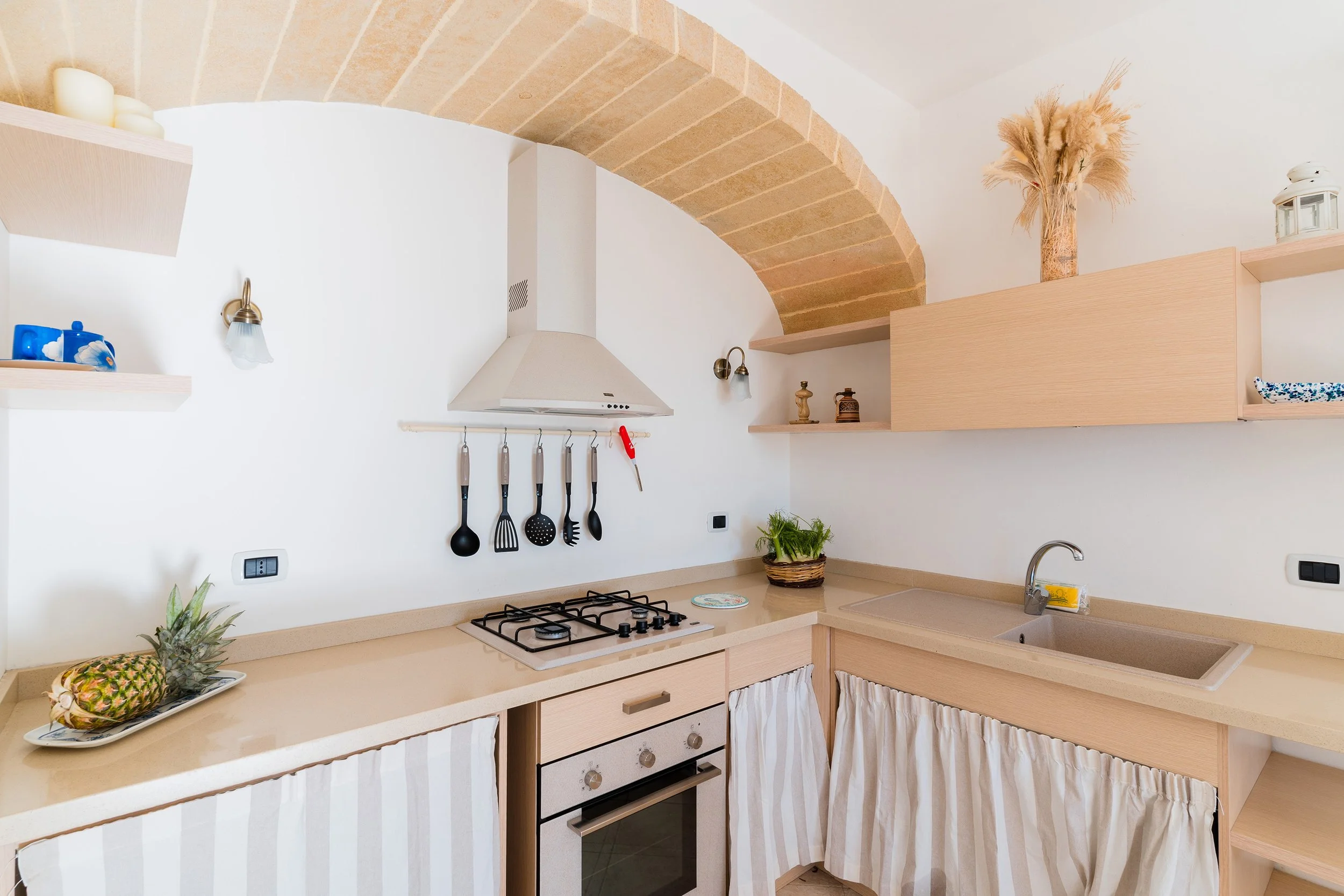 A kitchen with beige countertops, a stove, a pineapples on a tray, open shelving, a vase with dried plants, and some decorative items on the shelves. The ceiling has an arch of bricks.