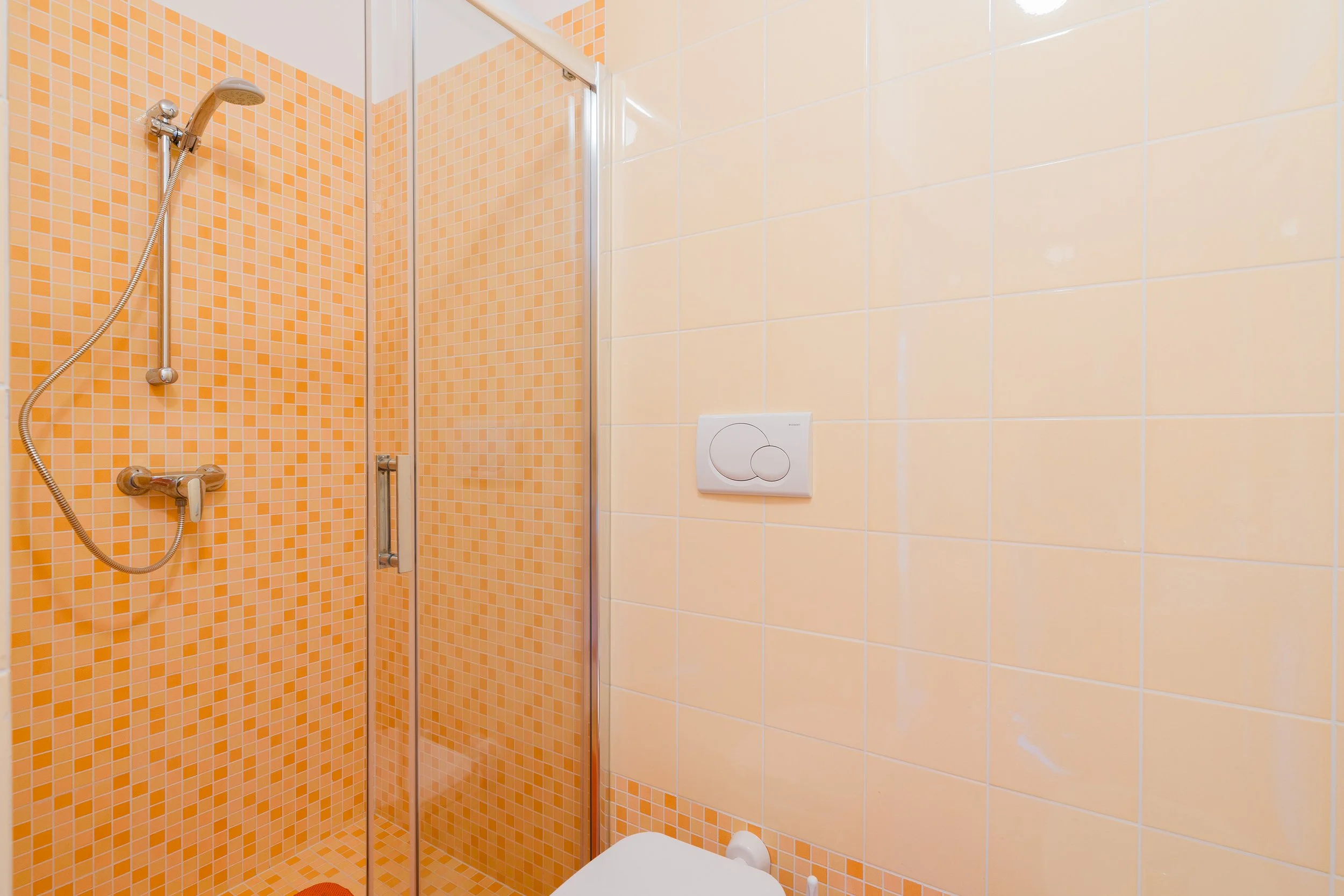Bathroom with orange and cream ceramic tiles, walk-in shower with glass door, showerhead, and controls, and a white wall-mounted toilet flush button.