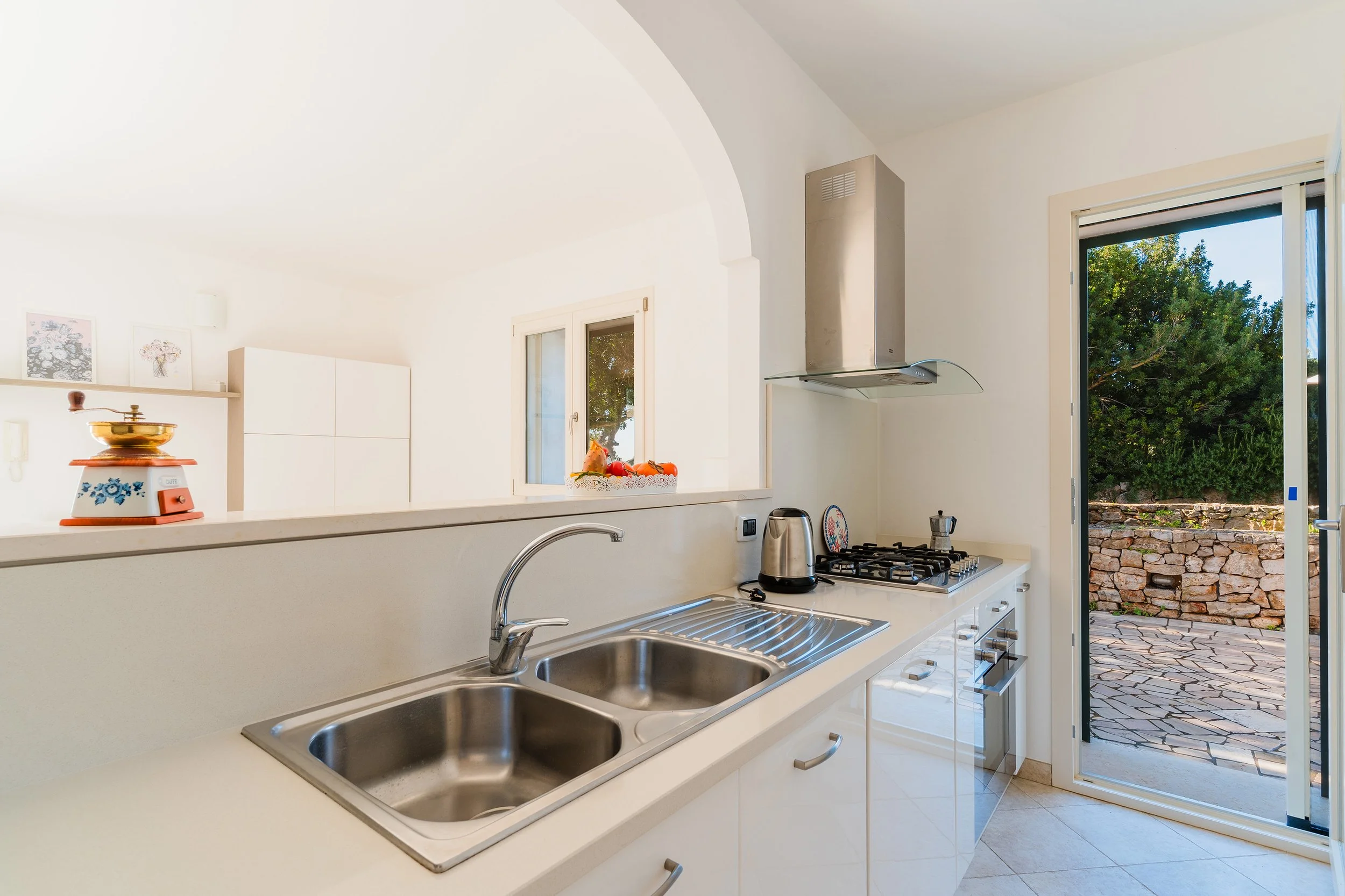 A bright kitchen with white cabinets, stainless steel sink, gas stove, and a stainless steel range hood. There is an open door leading outside to a patio with a stone wall and greenery. A small window displays trees outside.