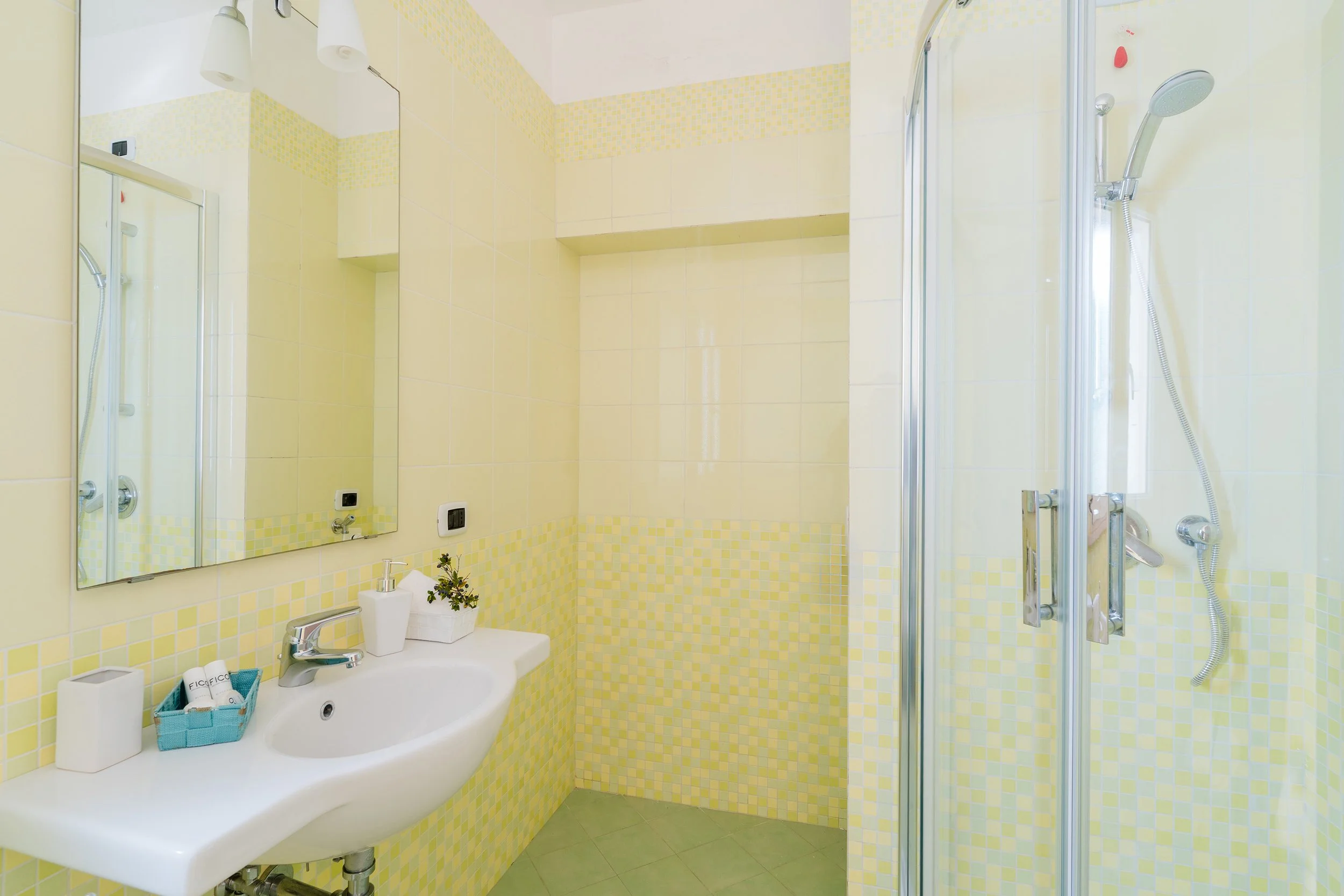 A small bathroom with yellow square mosaic tile accents, a white sink with a mirror above it, a small blue basket with toiletries, and a glass shower enclosure with a showerhead and red soap holder.