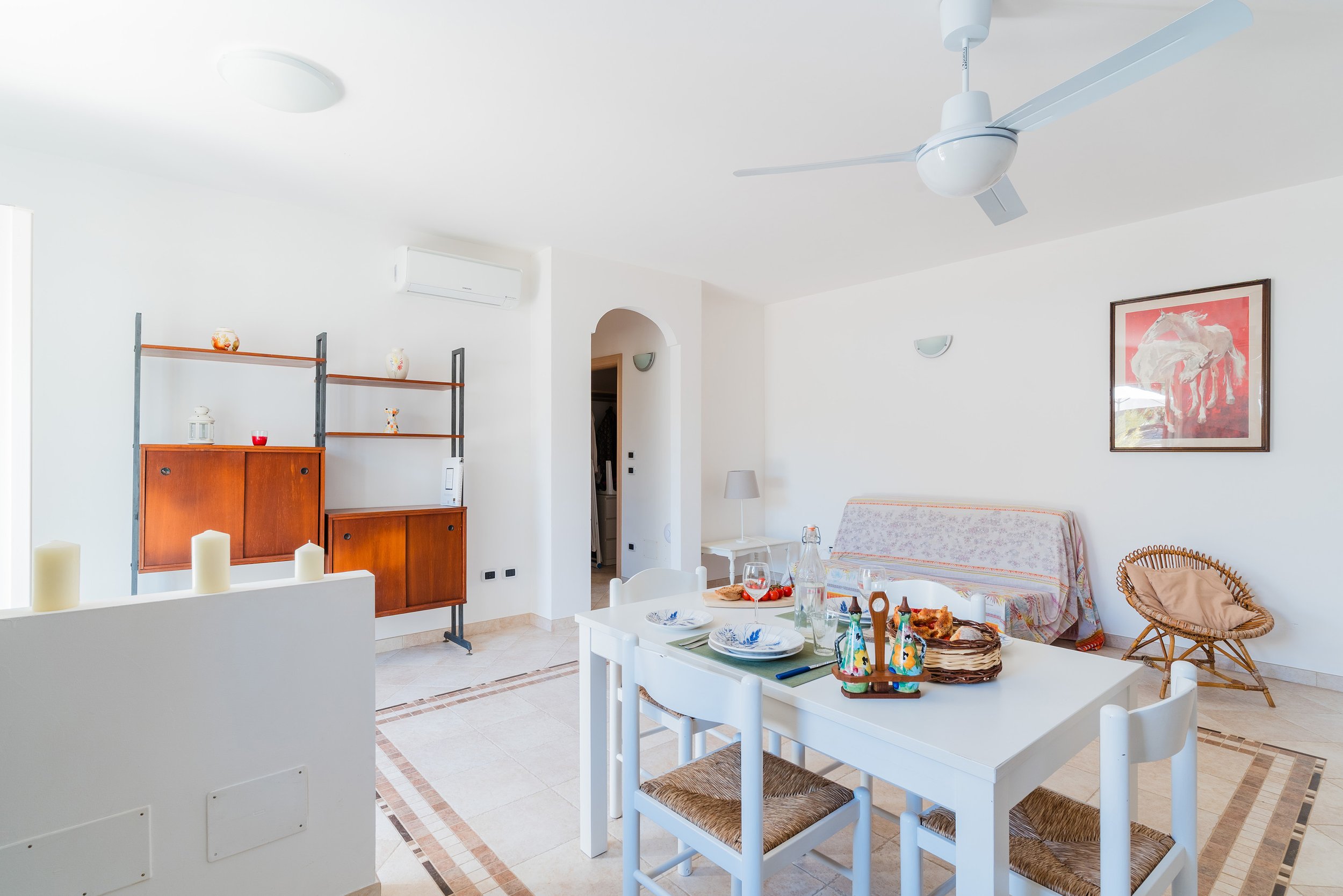 A bright living room with white walls, a white ceiling fan, and a framed artwork of a horse. There is a table set for a meal with plates, glasses, and food, and surrounding chairs with woven seats. A wooden shelving unit with decorative items is agai