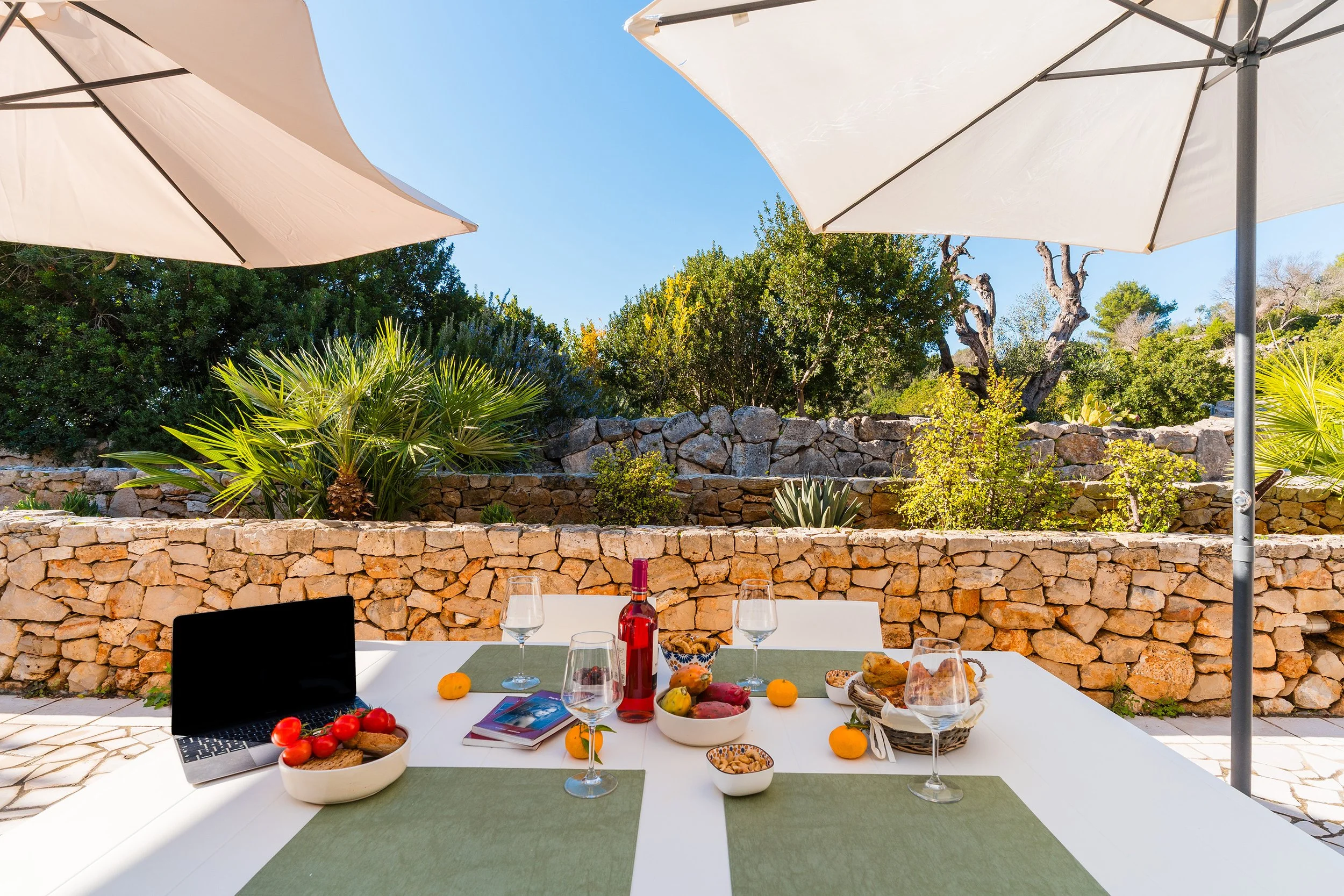 Outdoor dining table set with food and drinks, with white umbrellas and a landscaped backyard with trees and stone wall in the background.
