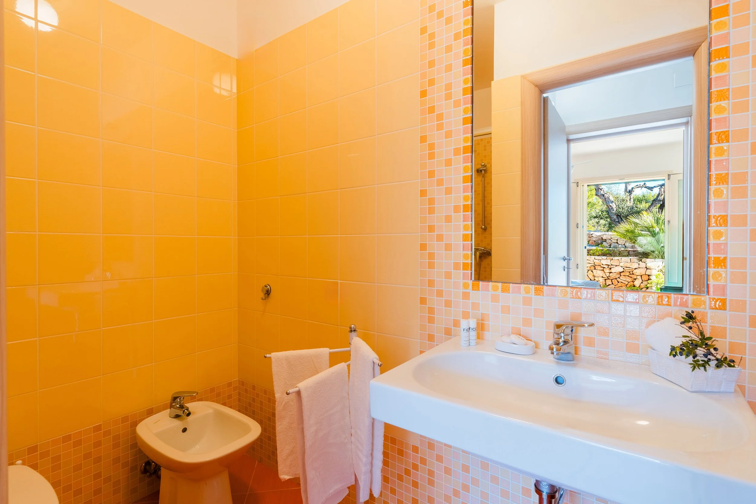 Bathroom with yellow tiles on walls, a mirror above the sink, a window showing outdoor greenery, and bathroom accessories like towels and toiletries.