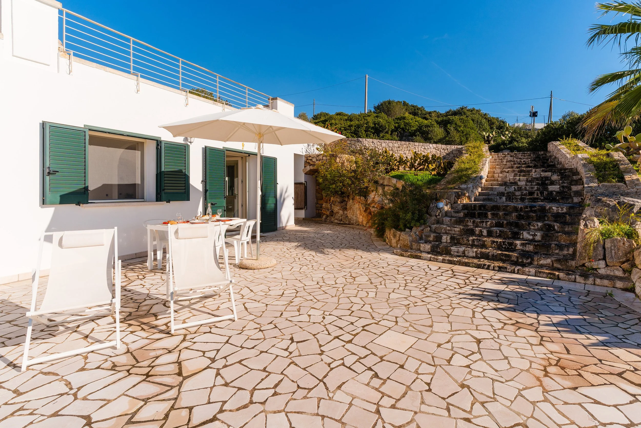 A patio with white patterned stone tiles, a white building with open green shutters, a white dining table with chairs and a large white umbrella, stone stairs, lush greenery, and a clear blue sky.