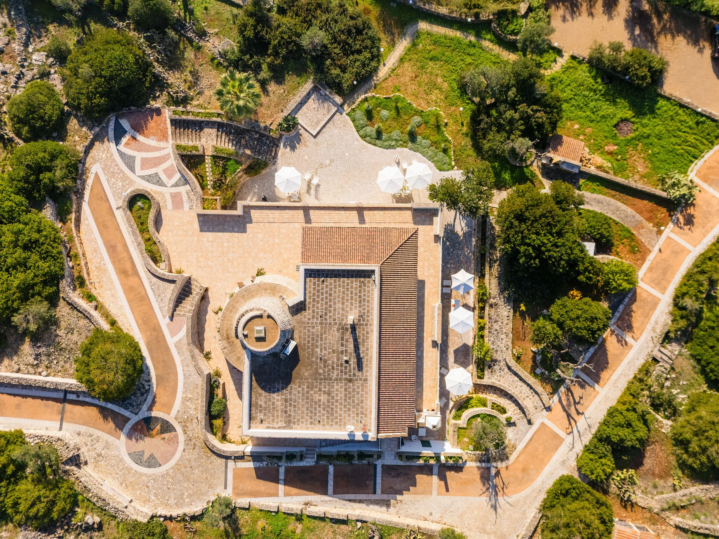 An aerial view of a lush, green garden with pathways, trees, and shrubbery surrounding a structure with a tiled roof. There are white umbrellas on a patio area.