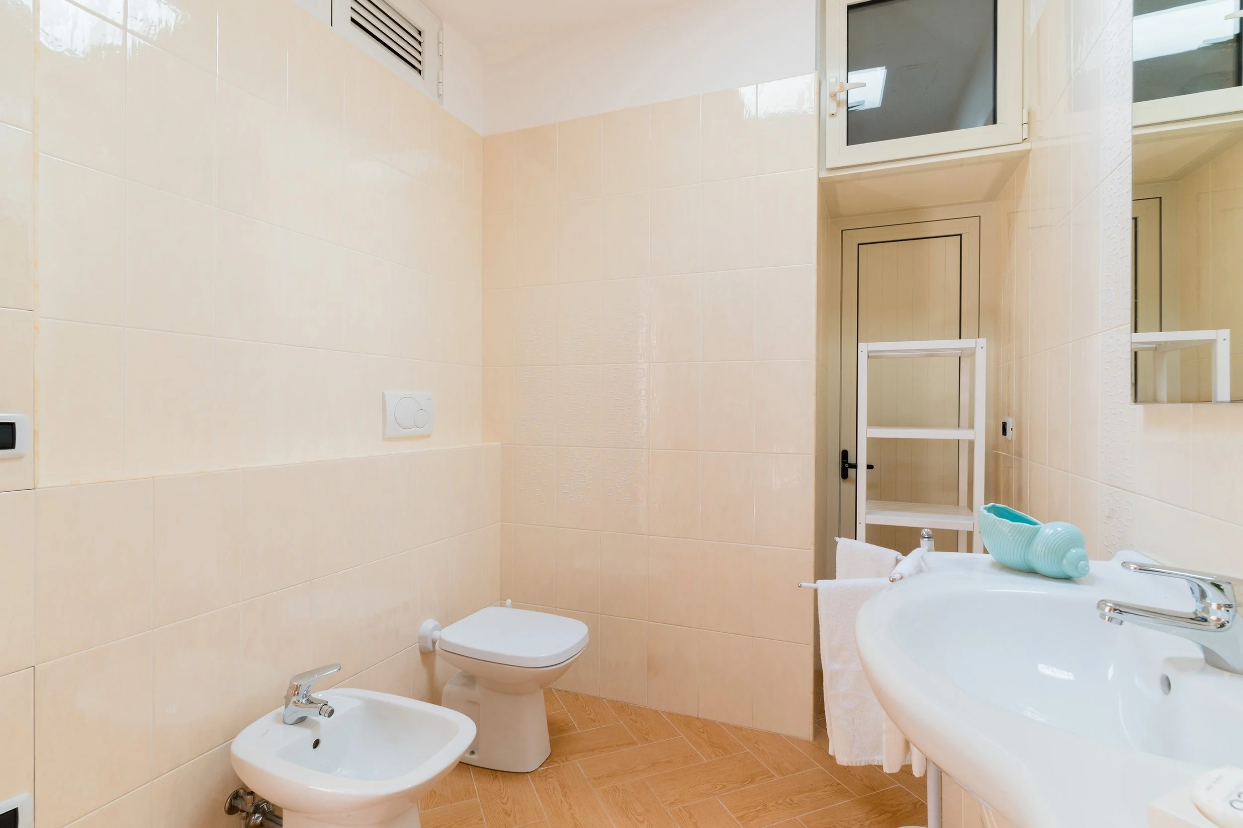A bathroom with beige tiled walls and wood-look tile flooring. It contains a white toilet, bidet, and an oval sink with a mirror above it. There is a small window above the toilet and bidet, a white shelving unit, and some towels and toiletries on th