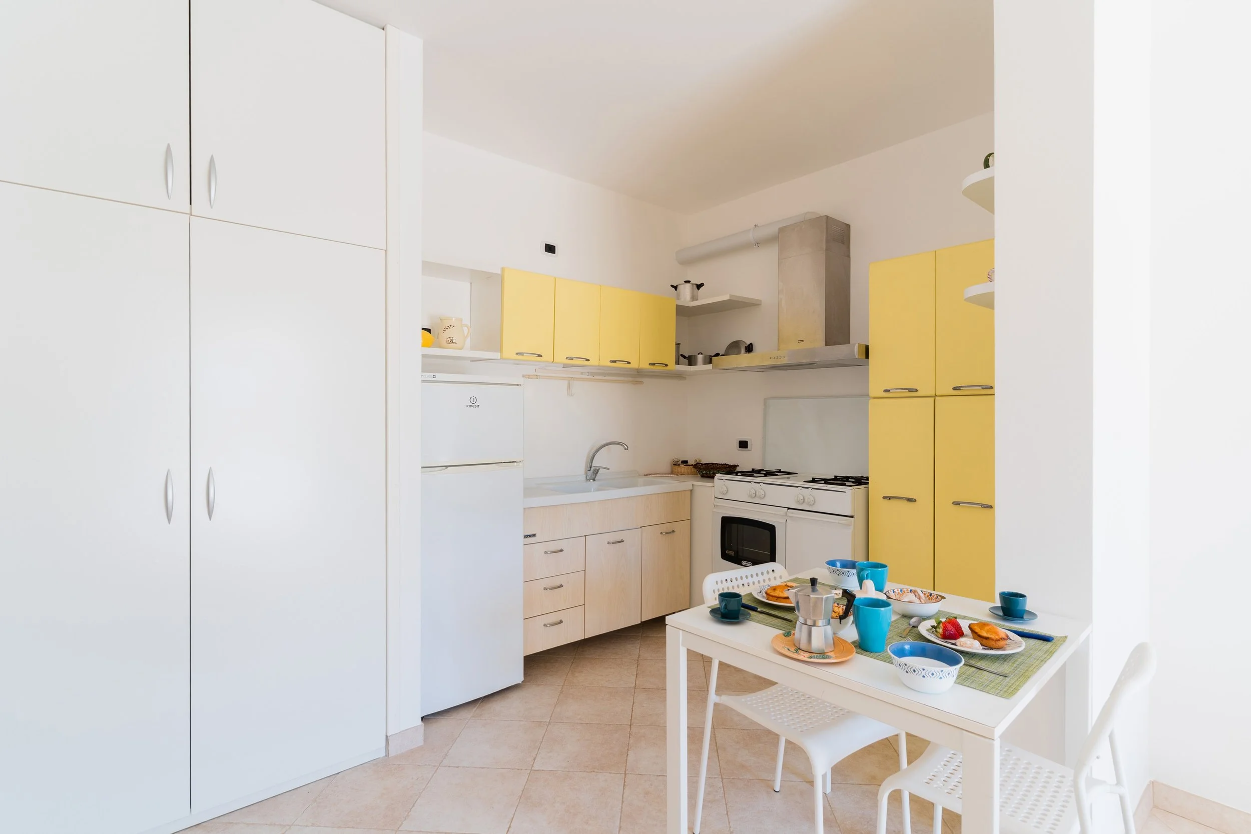 A cozy kitchen with yellow and white cabinets, a table set for a meal with cups, bowls, and plates, and a small white chair.