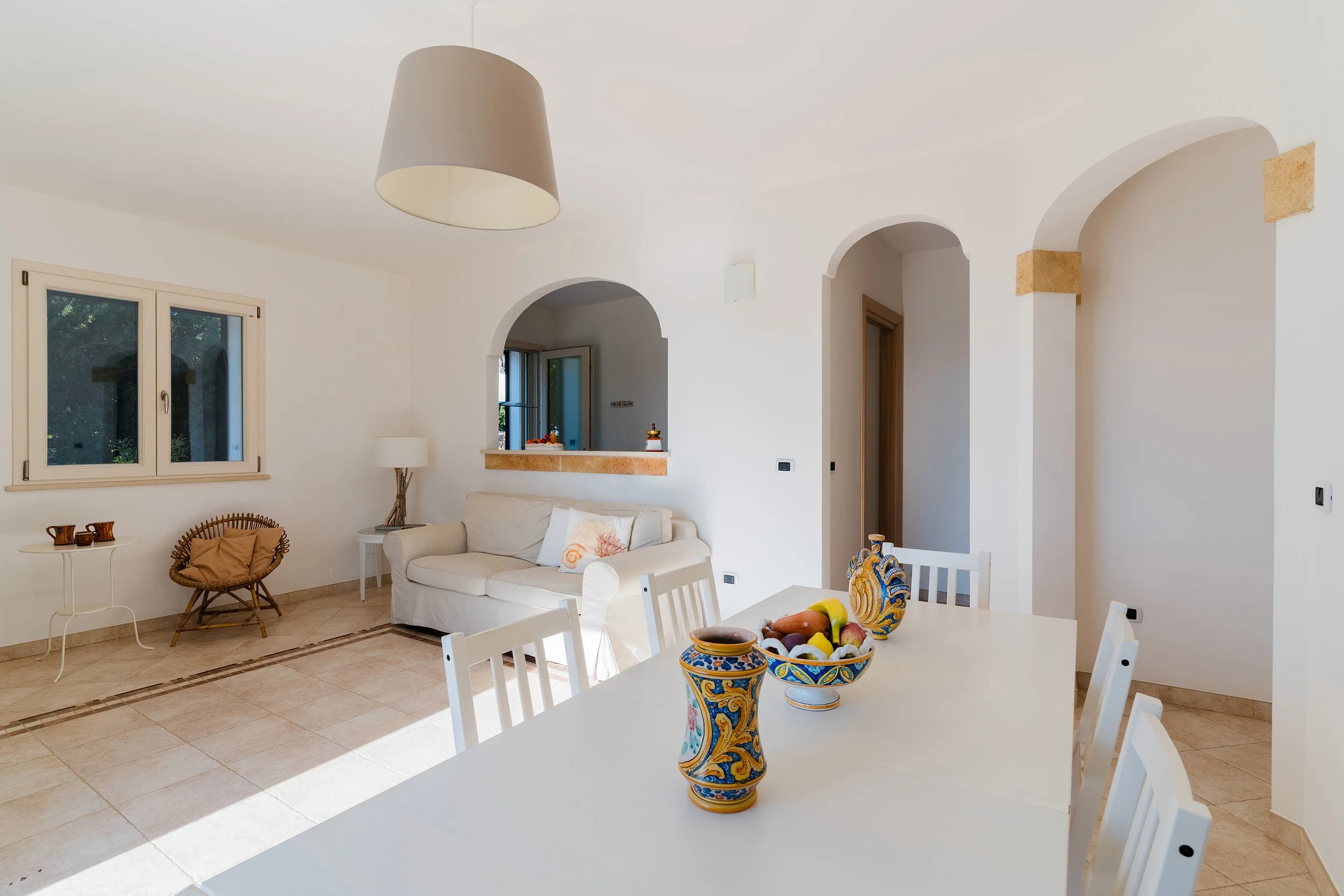A bright, minimalist living room with white walls, arched doorways, and beige tiled floors. The space includes a white couch with pillows, a white side table with a lamp, a wicker chair with a blanket, windows with views of greenery, and a dining tab
